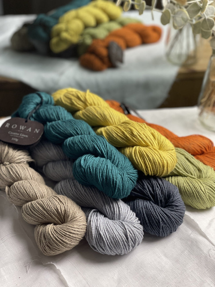 A selection of skeins of Rowan Creative Linen yarns on a white cloth with a mirror image in the background.
