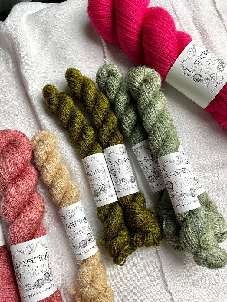A selection of skein of Highland 4Ply yarn in shades of green and pink and branded Inspiring Yarns lie on a white linen background.