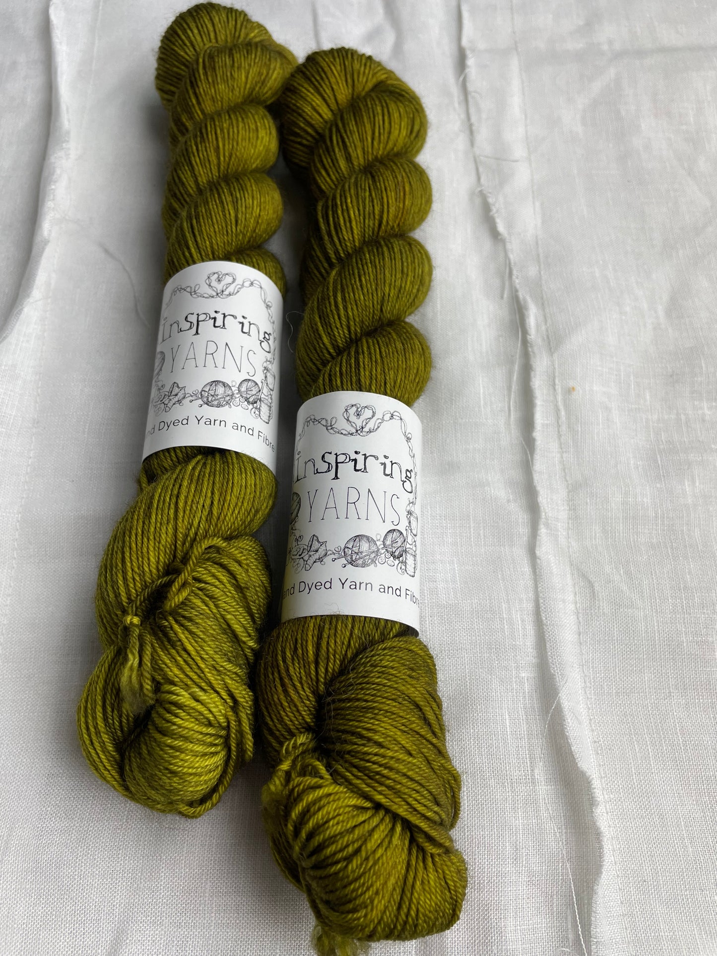 Two dark olive green coloured skeins of hand dyed Merino Smooth Sock yarn branded Inspiring Yarns on white linen fabric.