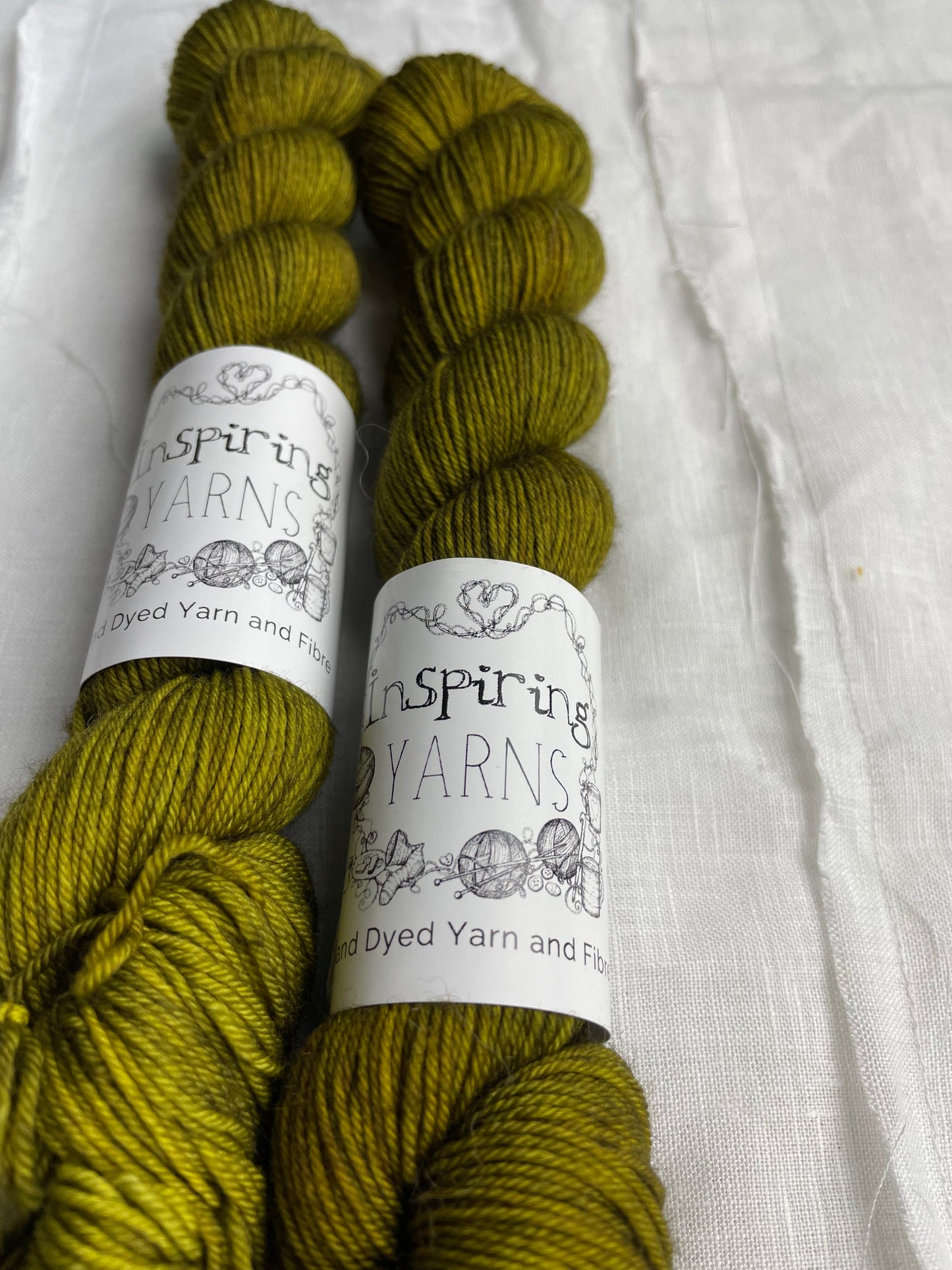 Close up image of two dark olive green coloured skeins of hand dyed Merino Smooth Sock yarn branded Inspiring Yarns on white linen fabric.