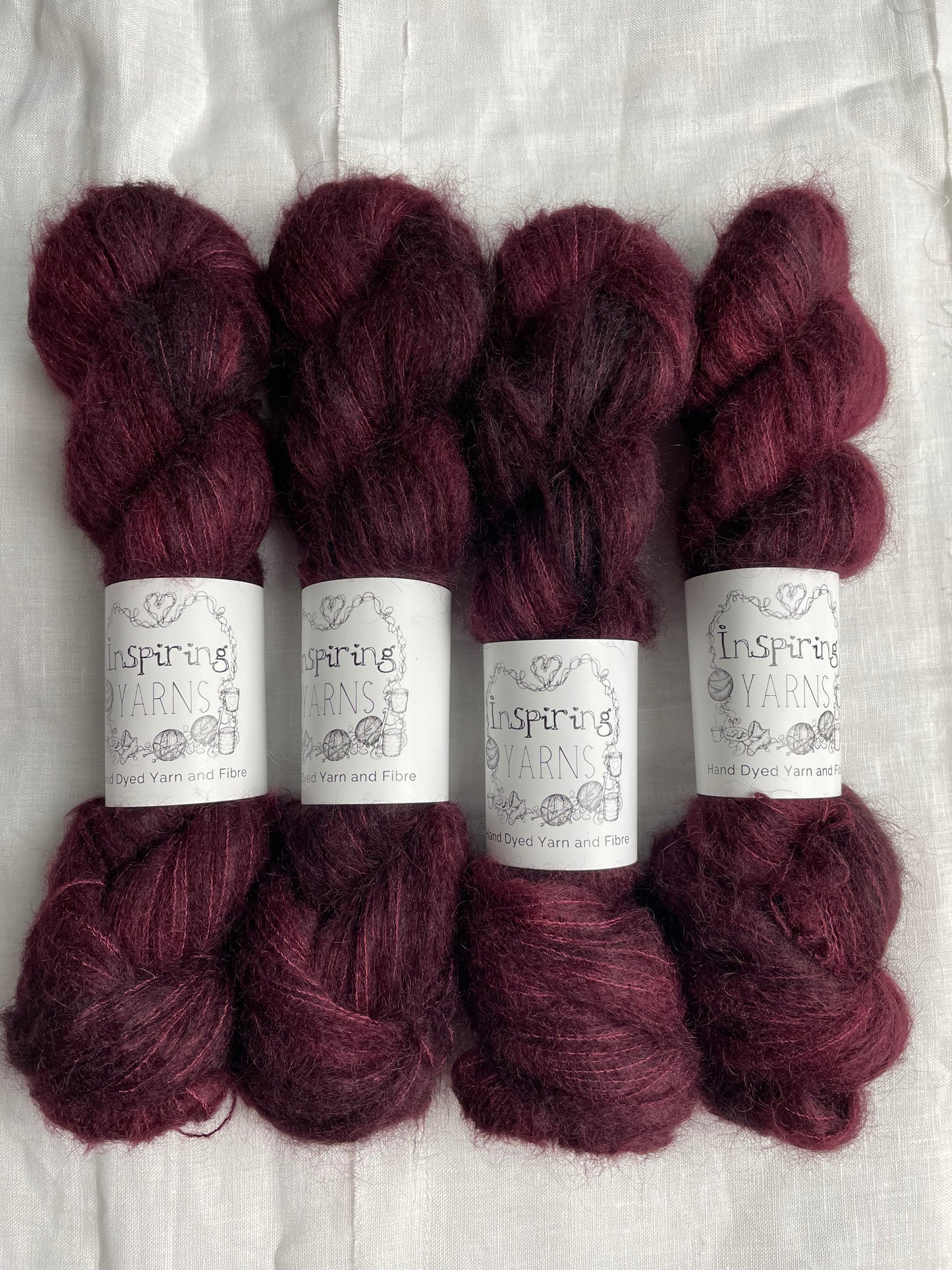 Four skeins of fluffy brushed suri silk lace yarn in a tonal dark burgundy colour labelled Inspiring Yarns on a white linen background.