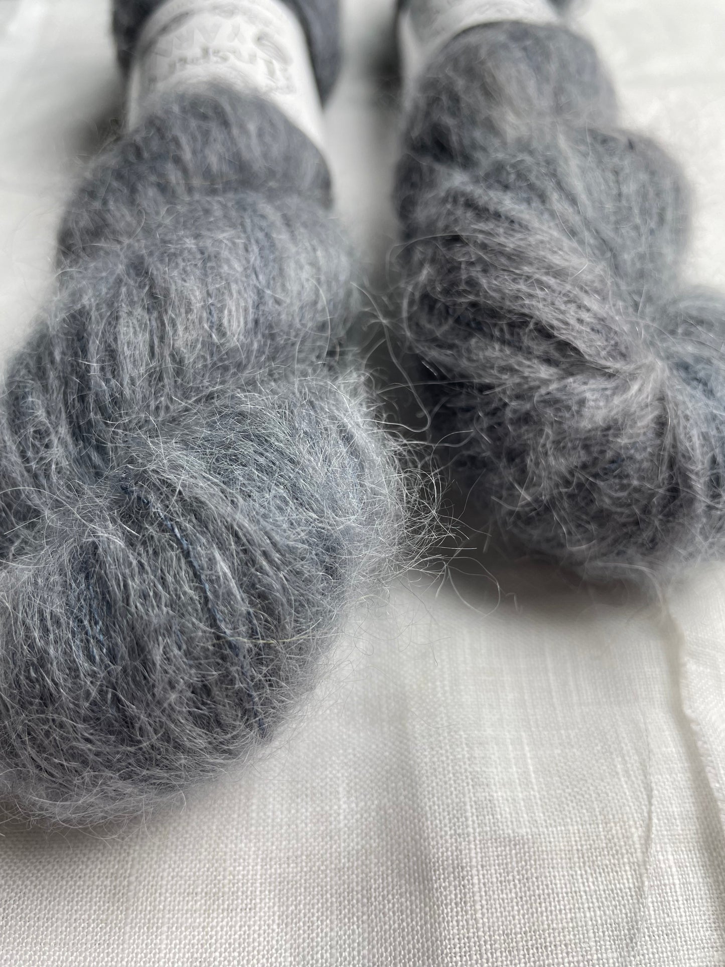 Close up of two skeins of tonal charcoal grey coloured hand dyed fluffy Alpaca Silk Lace yarn labelled Inspiring Yarns on a white linen background.