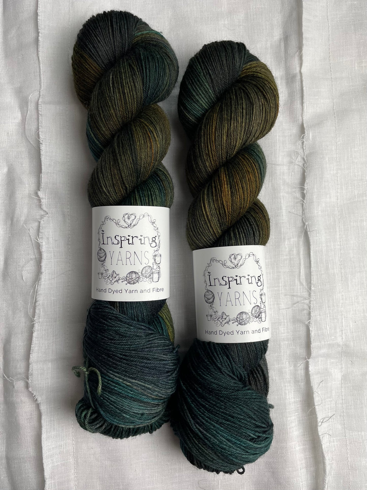 Two skeins of variegated black green gold and teal Merino Smooth Sock yarn labelled Inspiring Yarns on a white linen background