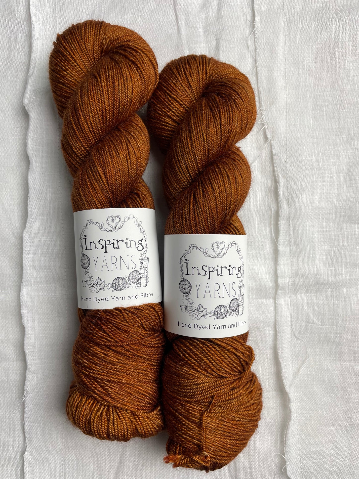 Two tonal burnt orange coloured skeins of hand dyed Yak Silk 4Ply yarn branded Inspiring Yarns on a white linen background.