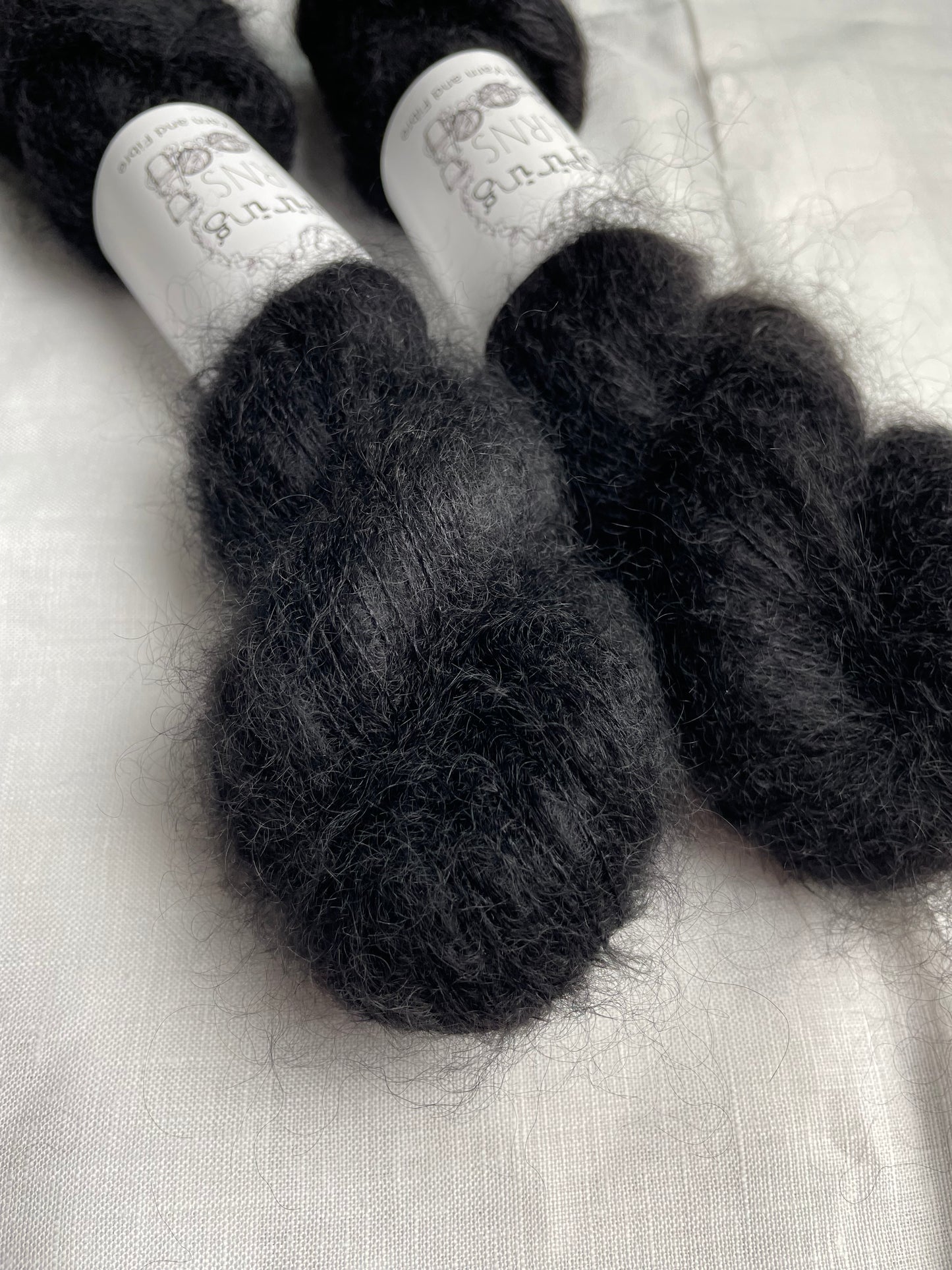 Two skeins of black coloured Kid Mohair Silk Lace labelled Inspiring Yarns on a white linen background.