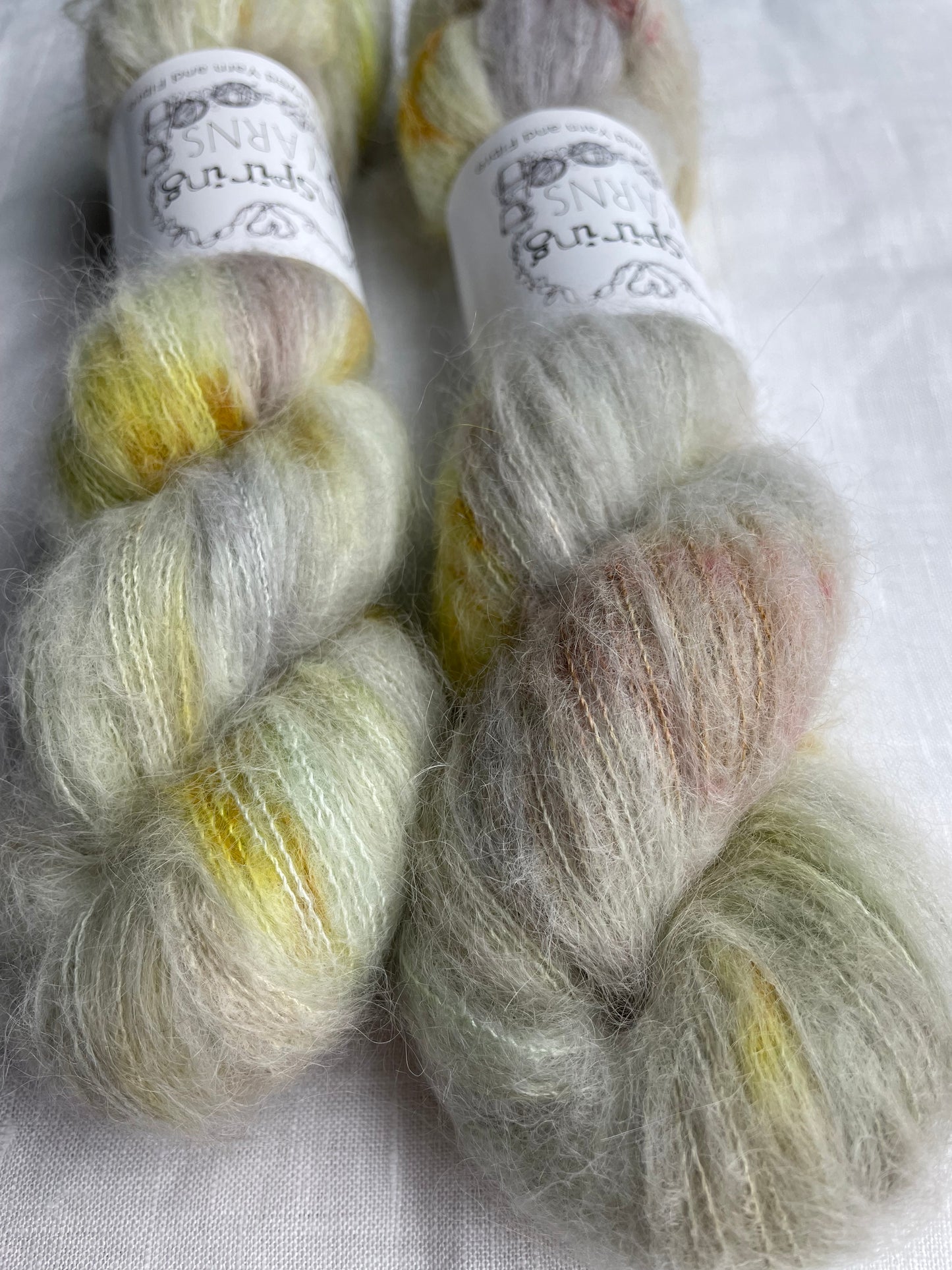 Close up of two skeins of speckled silver grey and mint coloured hand dyed fluffy Alpaca Silk Lace yarn labelled Inspiring Yarns on a white linen background.