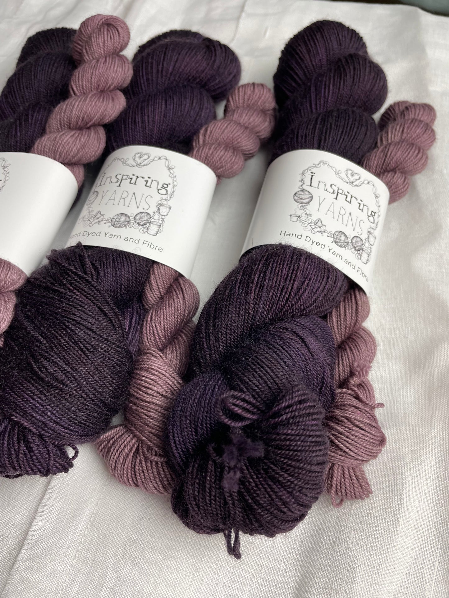 Close up of three sock sets made of a skein of dark tonal purple hand dyed yarn with a contrasting dusty lilac mini skein, labelled Inspiring Yarns and laid on a white linen cloth.