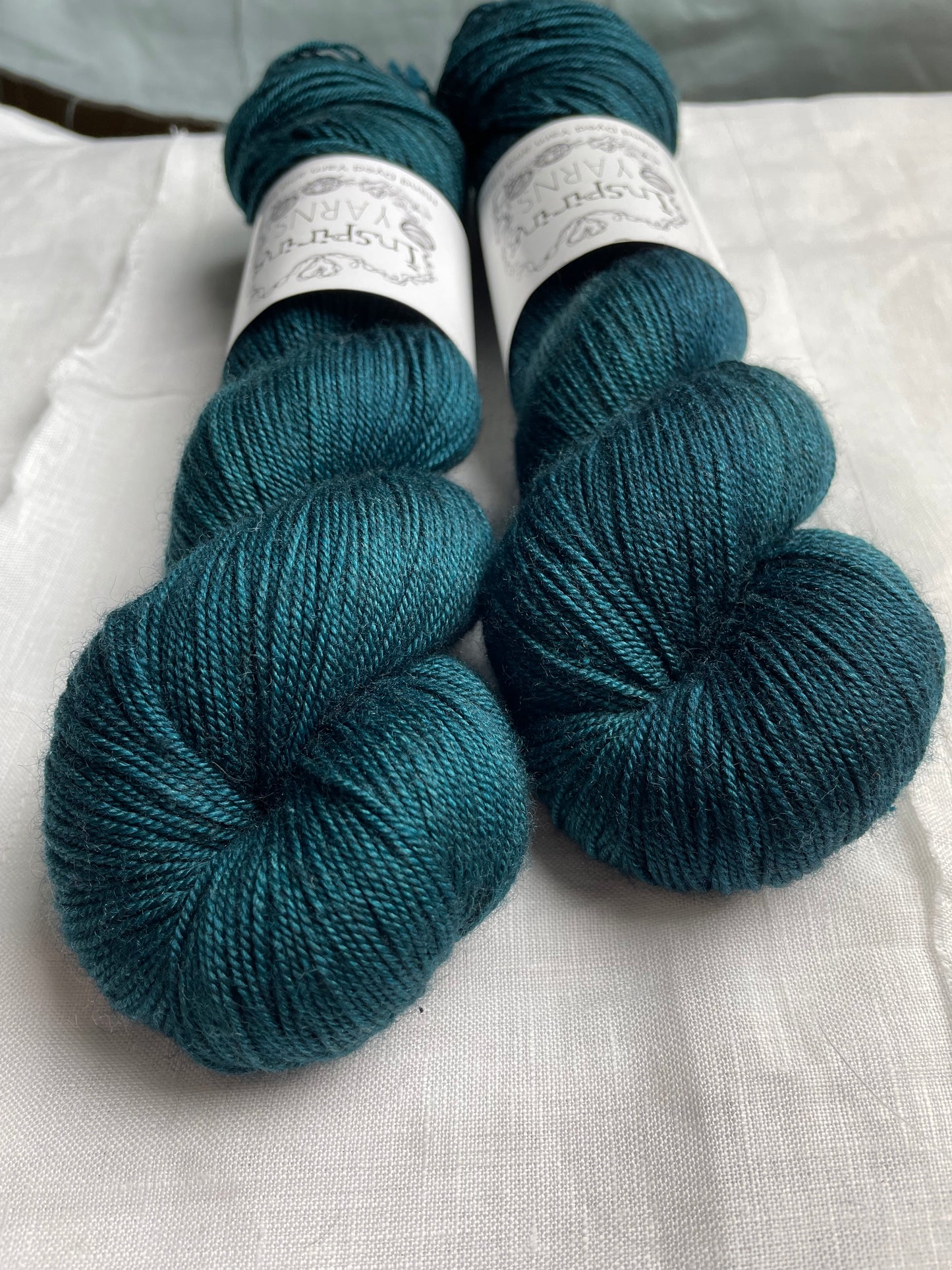 Close up of two tonal intense teal blue coloured skeins of hand dyed Yak Silk 4Ply yarn branded Inspiring Yarns on a white linen background.