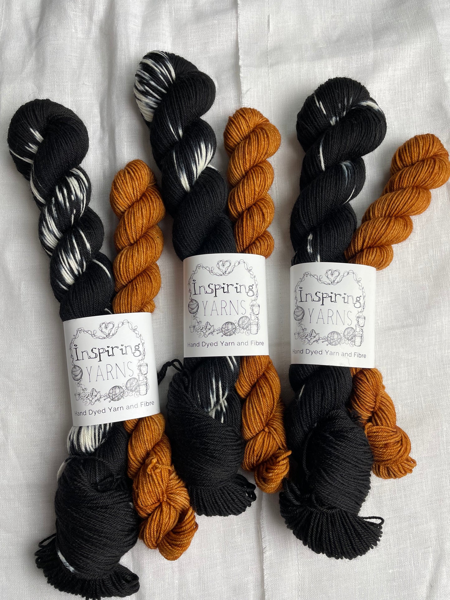 three sock sets made of a skein of black and white hand dyed yarn with a contrasting pumpkin orange mini skein, labelled Inspiring Yarns and laid on a white linen cloth.