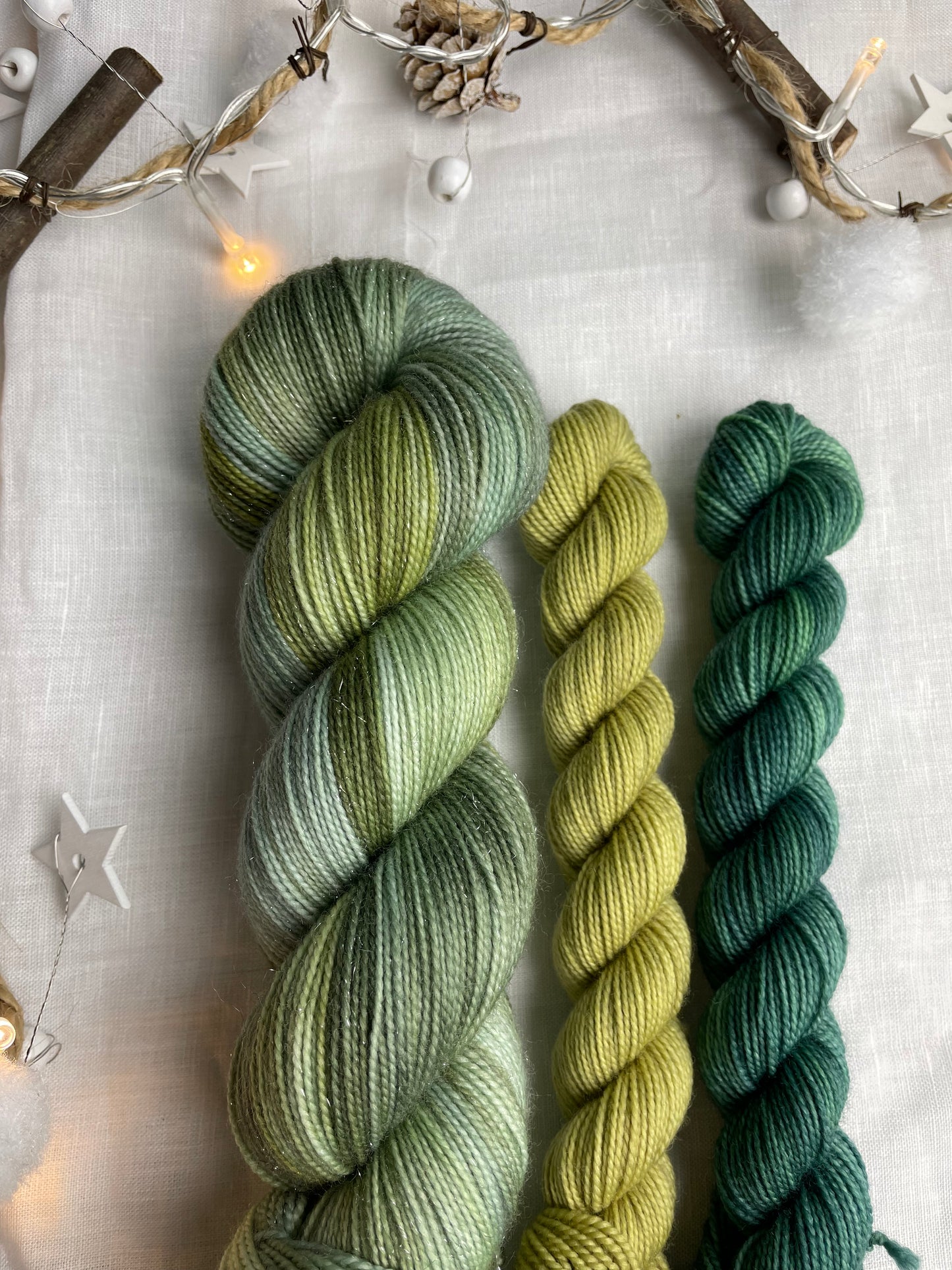 A hand dyed yarn sock set featuring large skein of Sparkle Sock Yarn in green tones with a teal and a lime green mini skein labelled Inspiring Yarns. The background is white linen and some Christmas lights with pinecones, twigs and stars.