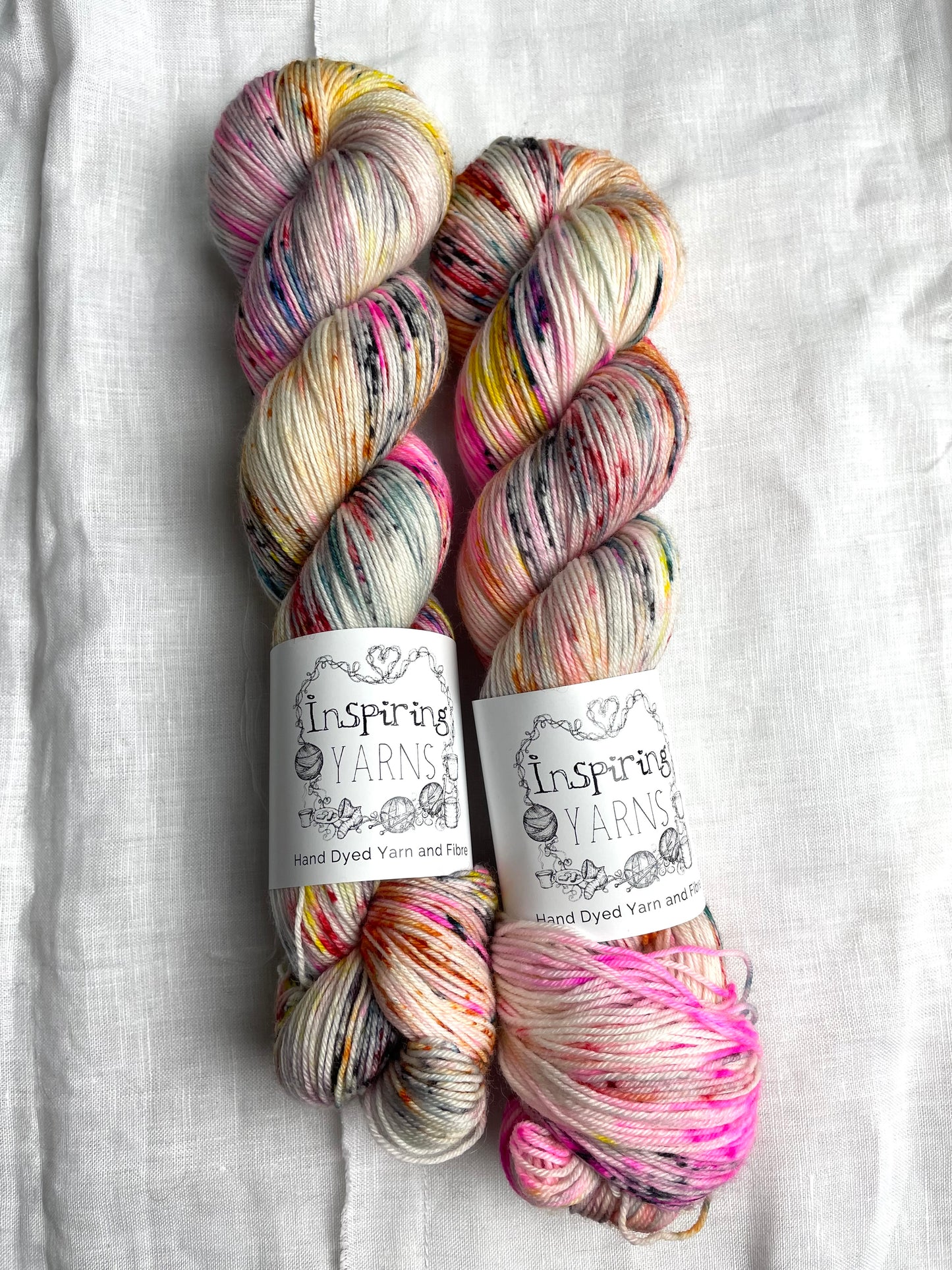 Two skeins of multi coloured speckled Merino Smooth Sock labelled InspiringYarns lies on a white linen background.
