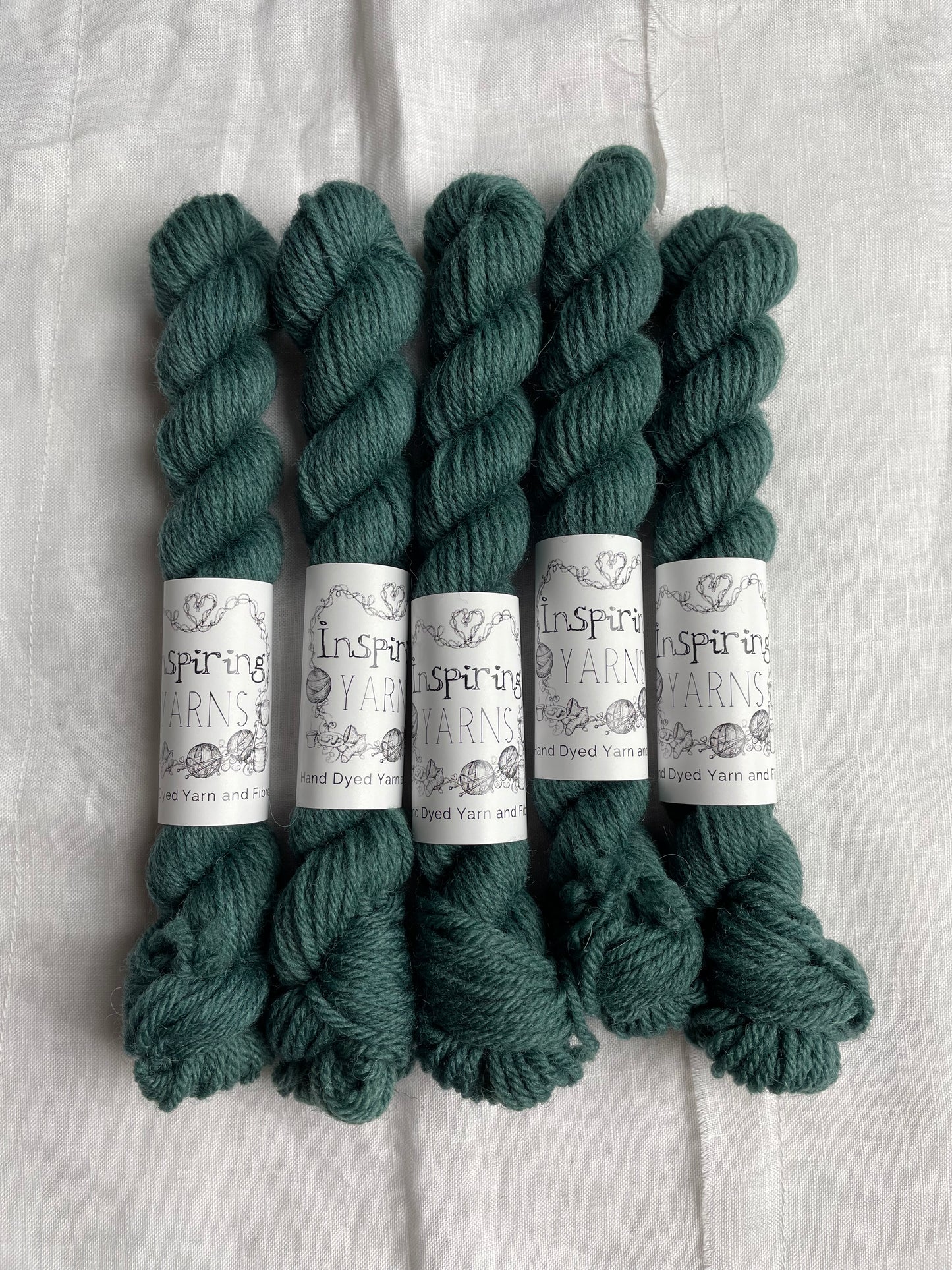 Five mini skeins of a tonal dark teal Highland DK yarn branded Inspiring Yarns on a white linen background.