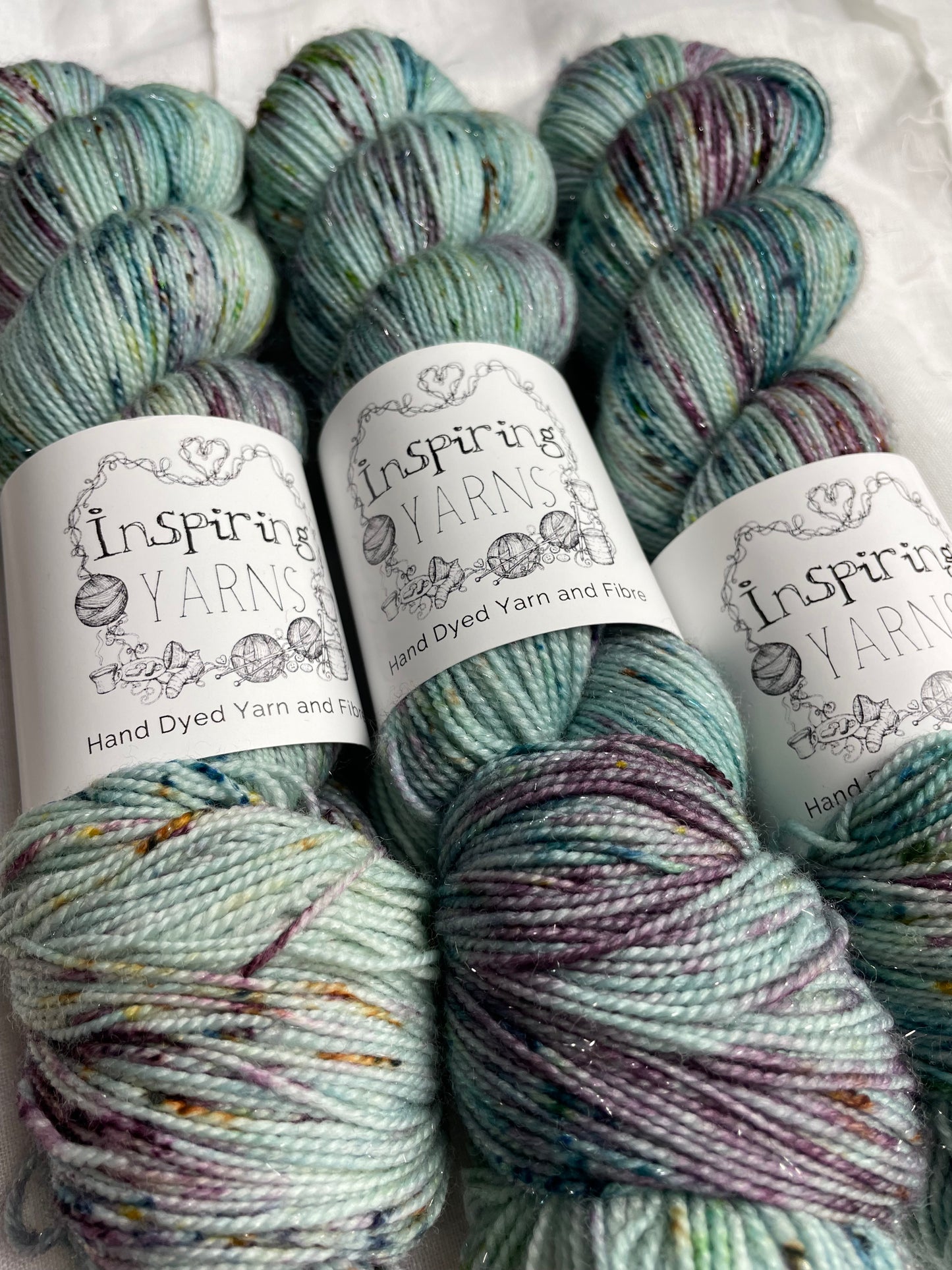 Three skeins of teal blue speckled Silver Sparkle Sock yarn labelled Inspiring Yarns on a white linen background.