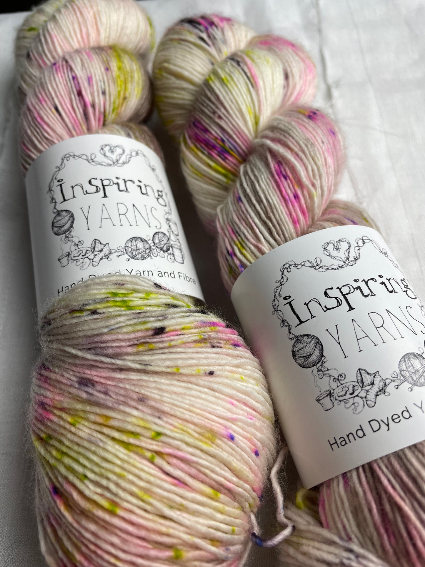 Close up of two skeins of speckled natural coloured hand dyed Merino Singles yarn labelled Inspiring Yarns on a white linen background.