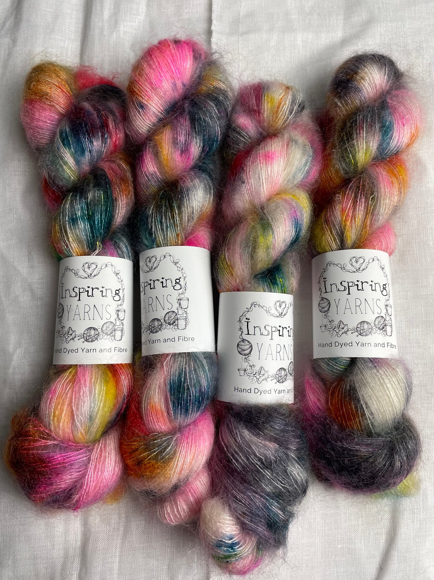 Four skeins of multi coloured speckled kid mohair silk lace yarn labelled Inspiring Yarns on a white linen background.