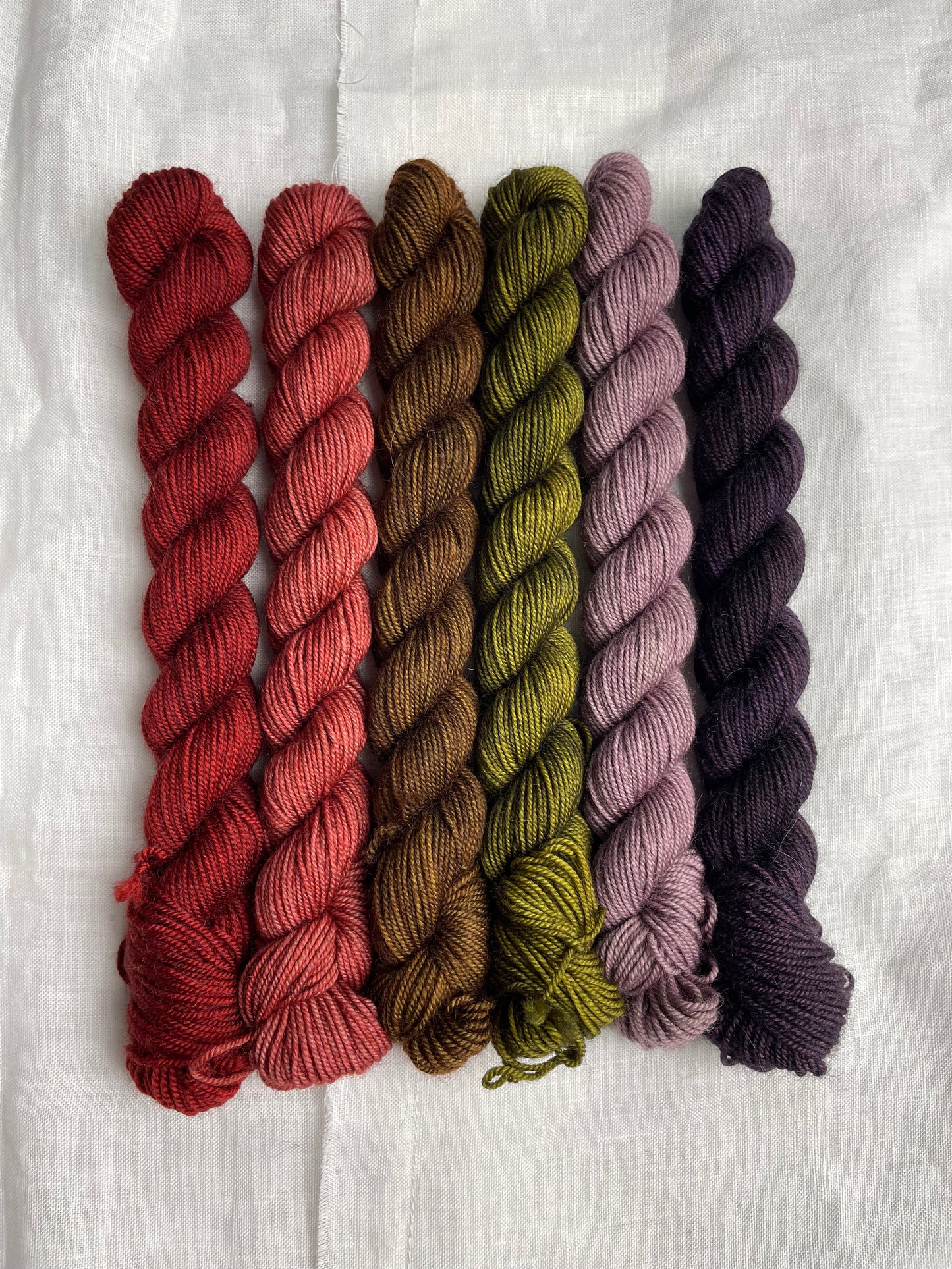 Close up of a set of 6 mini skeins of various colours of Yak Sock yarn on a white linen background.