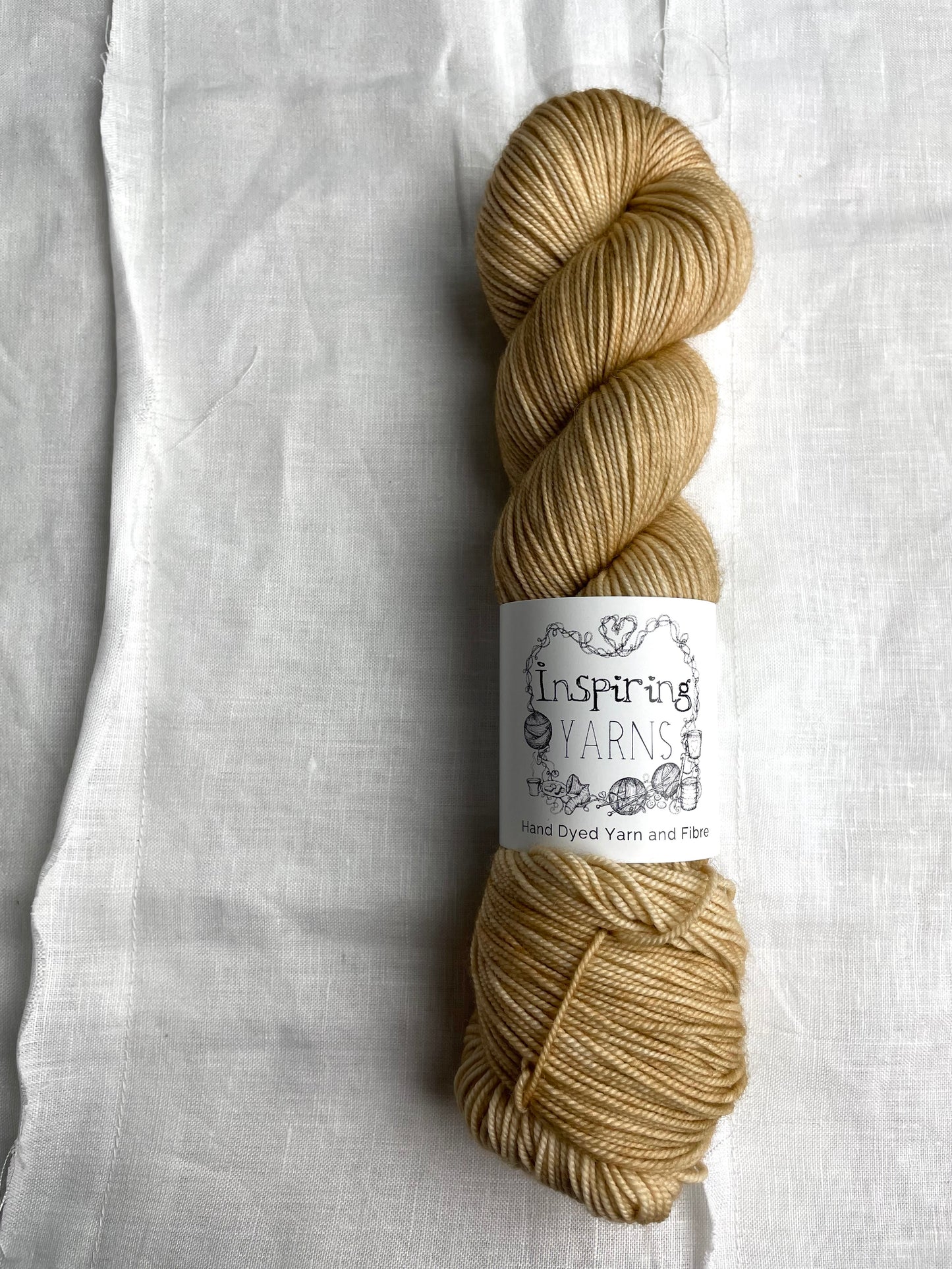 A skein of tonal golden cream coloured hand dyed Merino Sport yarn branded Inspiring Yarns on a background of white linen.
