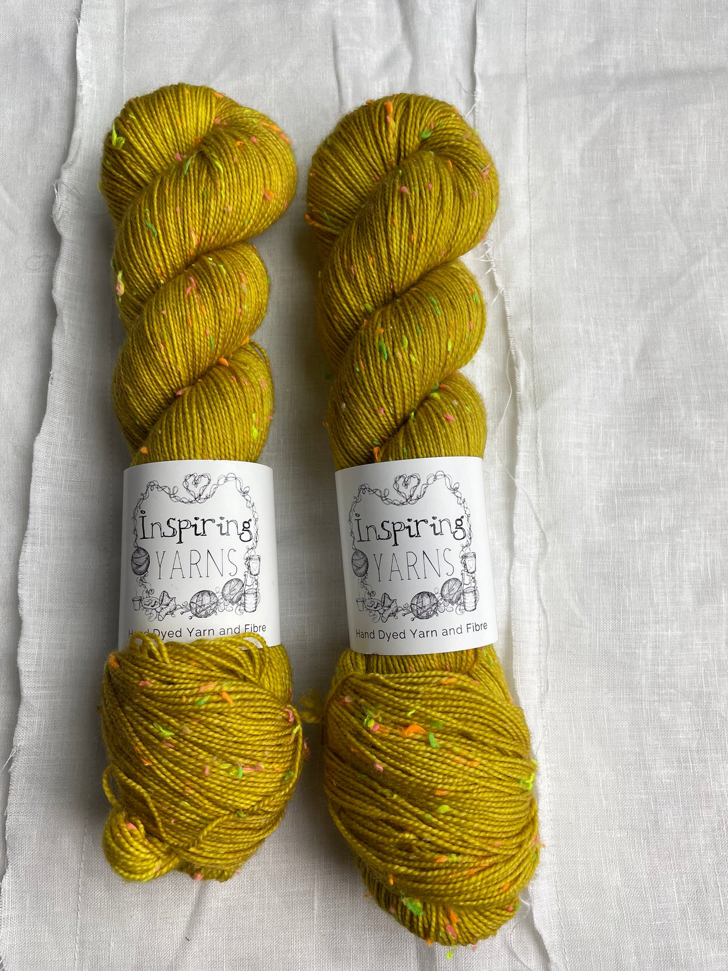 Two skeins of tonal golden ochre coloured hand dyed Disco Donegal yarn branded Inspiring Yarns on a white linen background. The yarn has multi coloured neon neps.