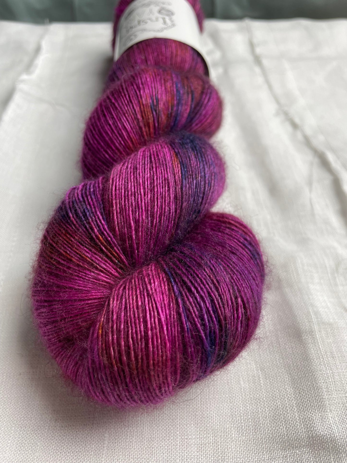 Close up of one speckled fuchsia pink coloured skeins of hand dyed Yak Silk Singles branded Inspiring Yarns on a white linen background.