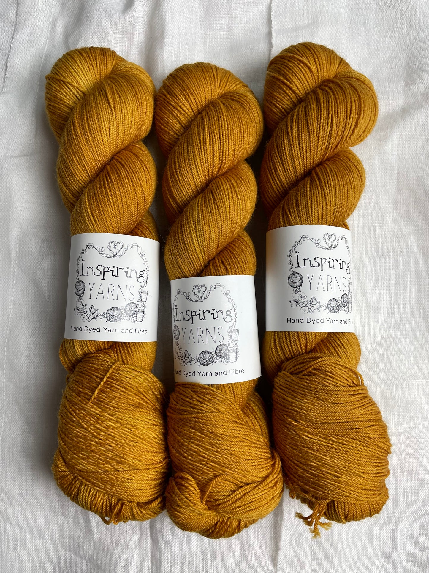 Three skeins of Keen As, a deep mustard tonal coloured Merino Smooth Sock, labelled Inspiring Yarns on a white linen background.