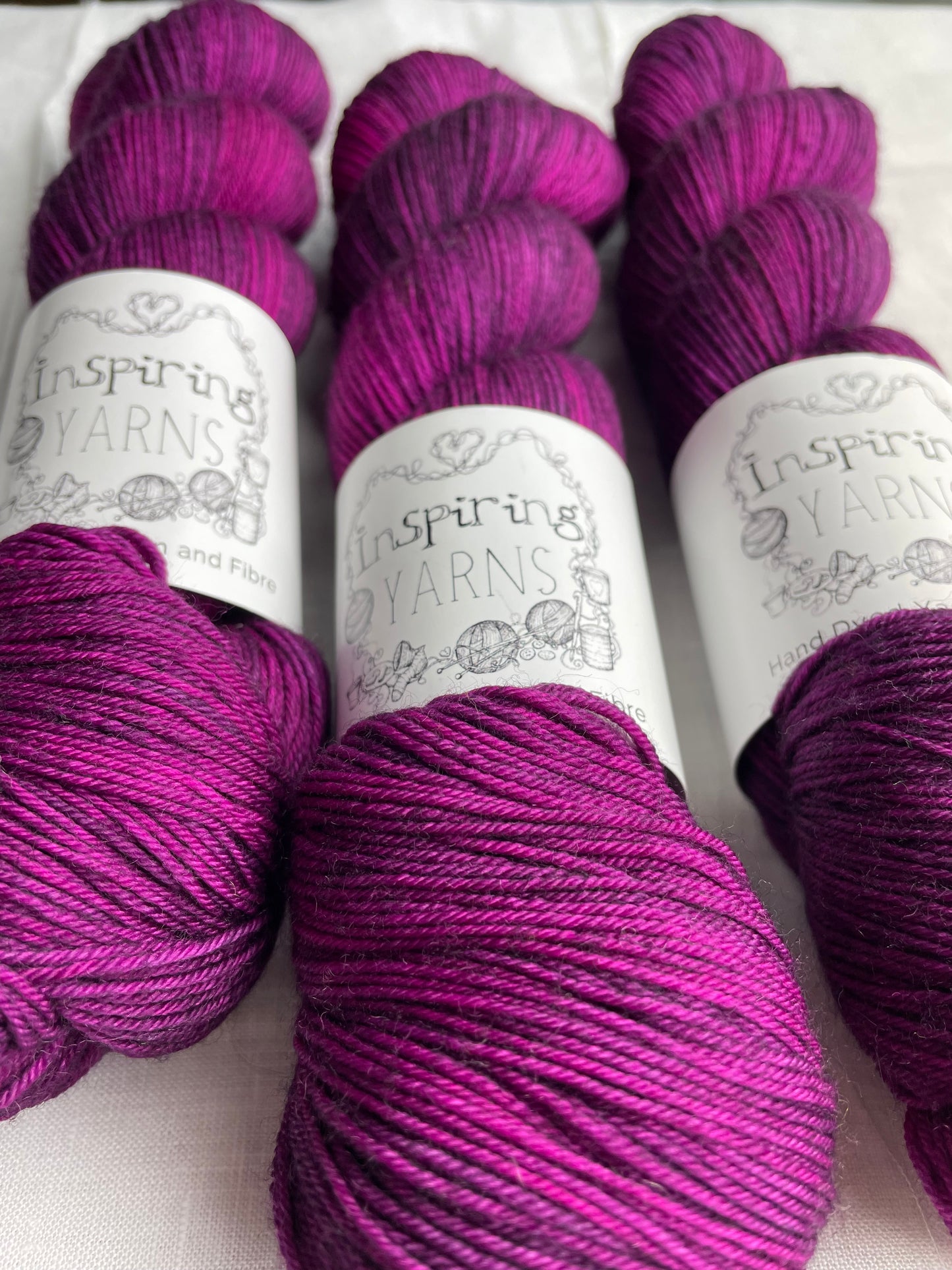 Close up of three lightly variegated cerise pink and dark purple coloured skeins of Merino Smooth Sock yarn branded Inspiring Yarns on a white linen fabric background.