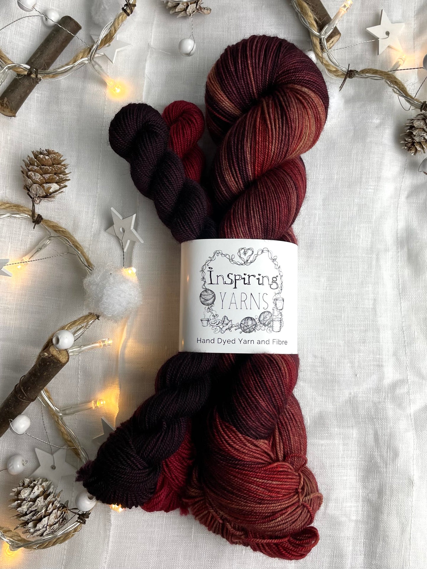 Hand dyed yarn sock set featuring large skein in burgundy, red and orange tones with a dark red and a burgundy mini skein labelled Inspiring Yarns. The background is white linen and some Christmas lights with pinecones, twigs and stars.