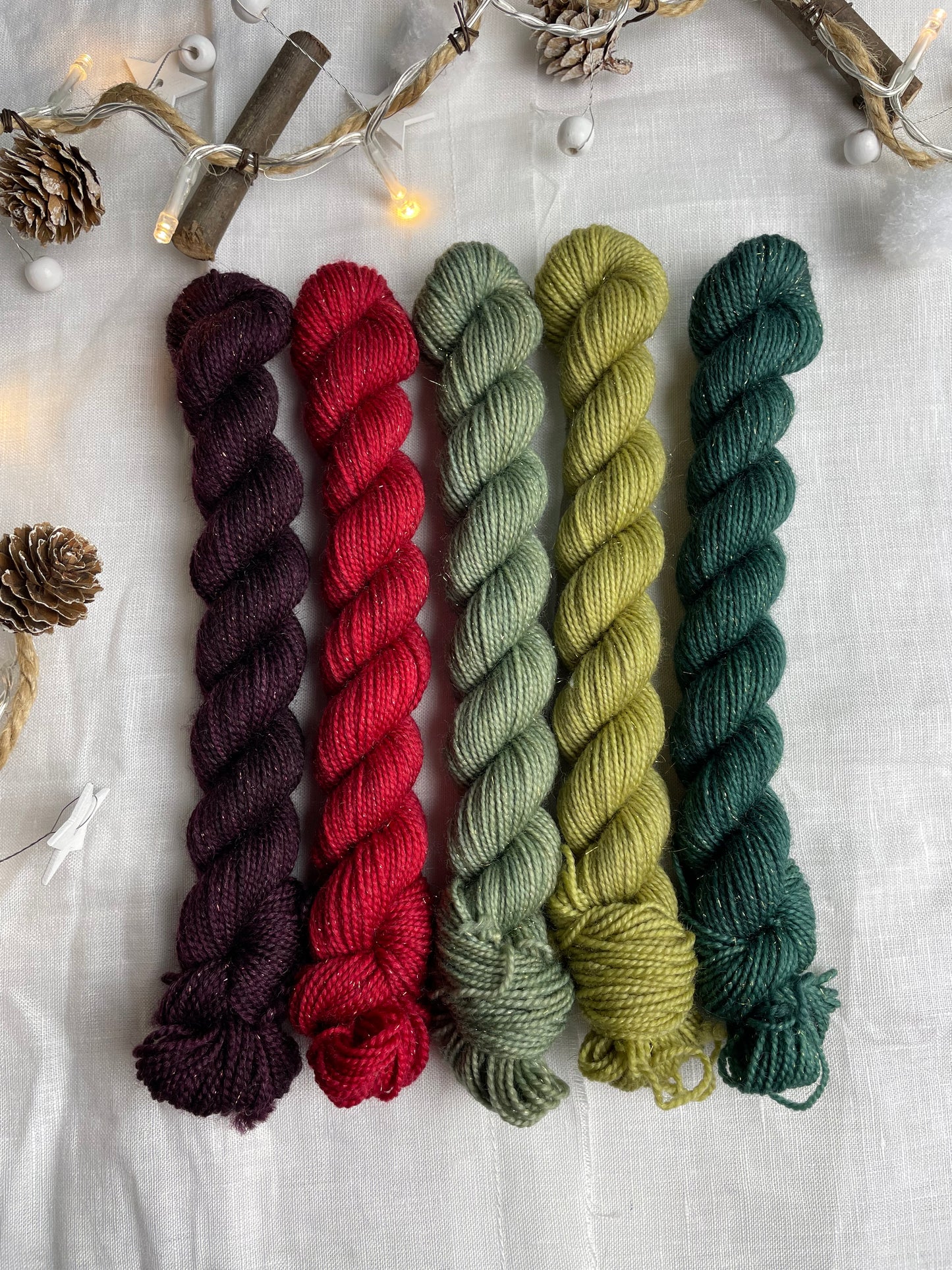 A set of five hand dyed yarn mini skeins on Sparkle Sock Yarn in red and green tones labelled Inspiring Yarns. The background is white linen and some Christmas lights with pinecones, twigs and stars.