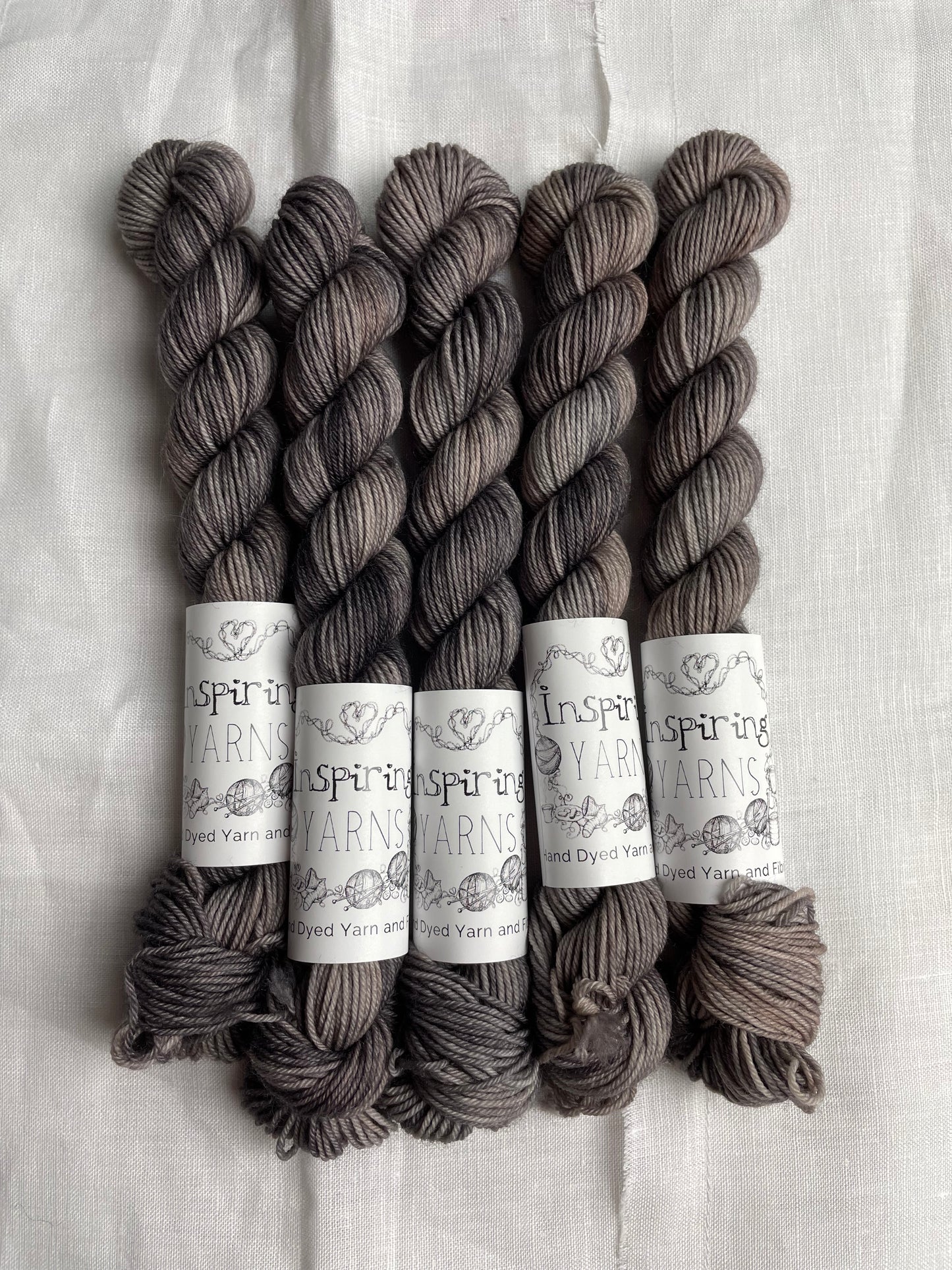 Five mini skeins of Merino Smooth Sock yarn hand dyed in a tonal grey colourway and labelled Inspiring Yarns on a white linen background.