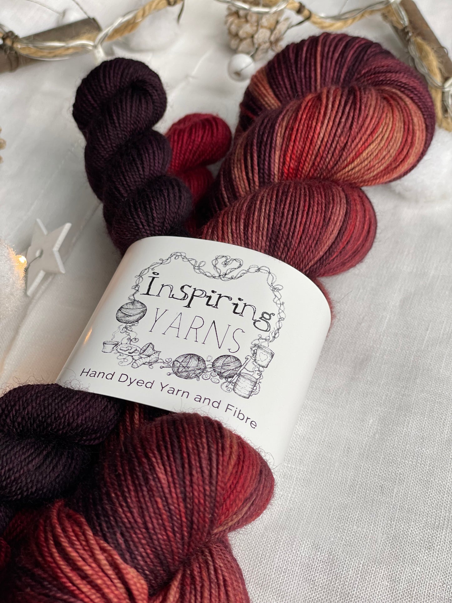Close up of a hand dyed yarn sock set featuring large skein in burgundy, red and orange tones with a dark red and a burgundy mini skein labelled Inspiring Yarns. The background is white linen and some Christmas lights with pinecones, twigs and stars.