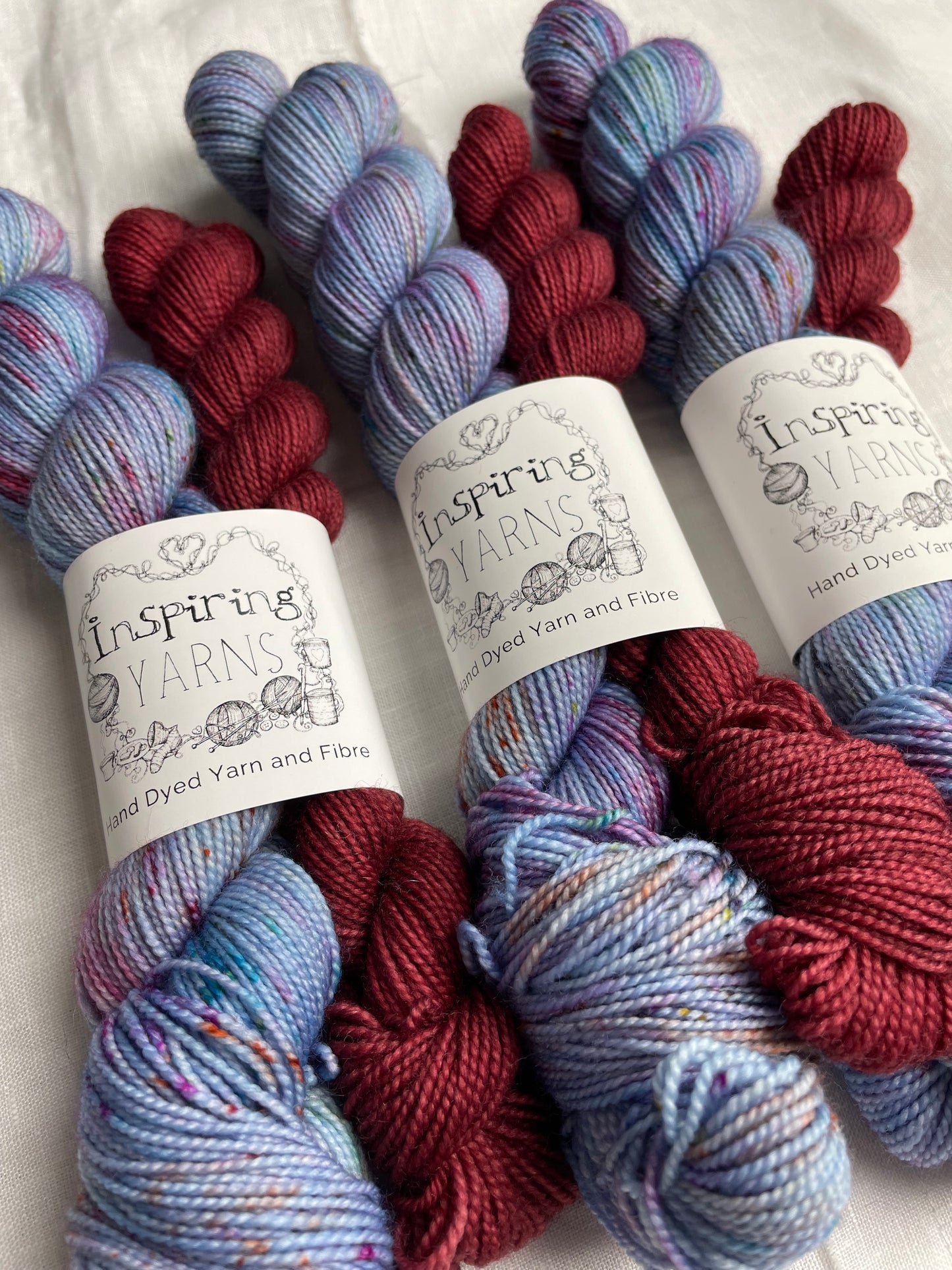 Close up of three sock sets made of a skein of light blue speckled hand dyed yarn with a contrasting deep blood red mini skein, labelled Inspiring Yarns and laid on a white linen cloth.