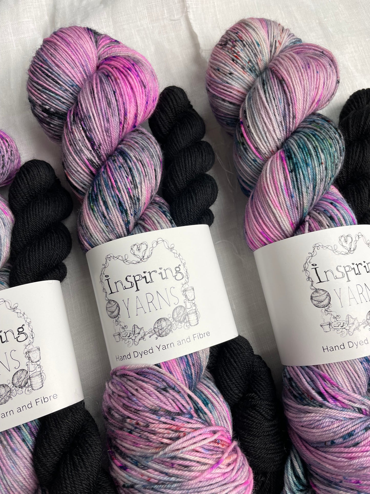 Close up of three sock sets made of a skein of pink black and teal speckled hand dyed yarn with a contrasting black mini skein, labelled Inspiring Yarns and laid on a white linen cloth.