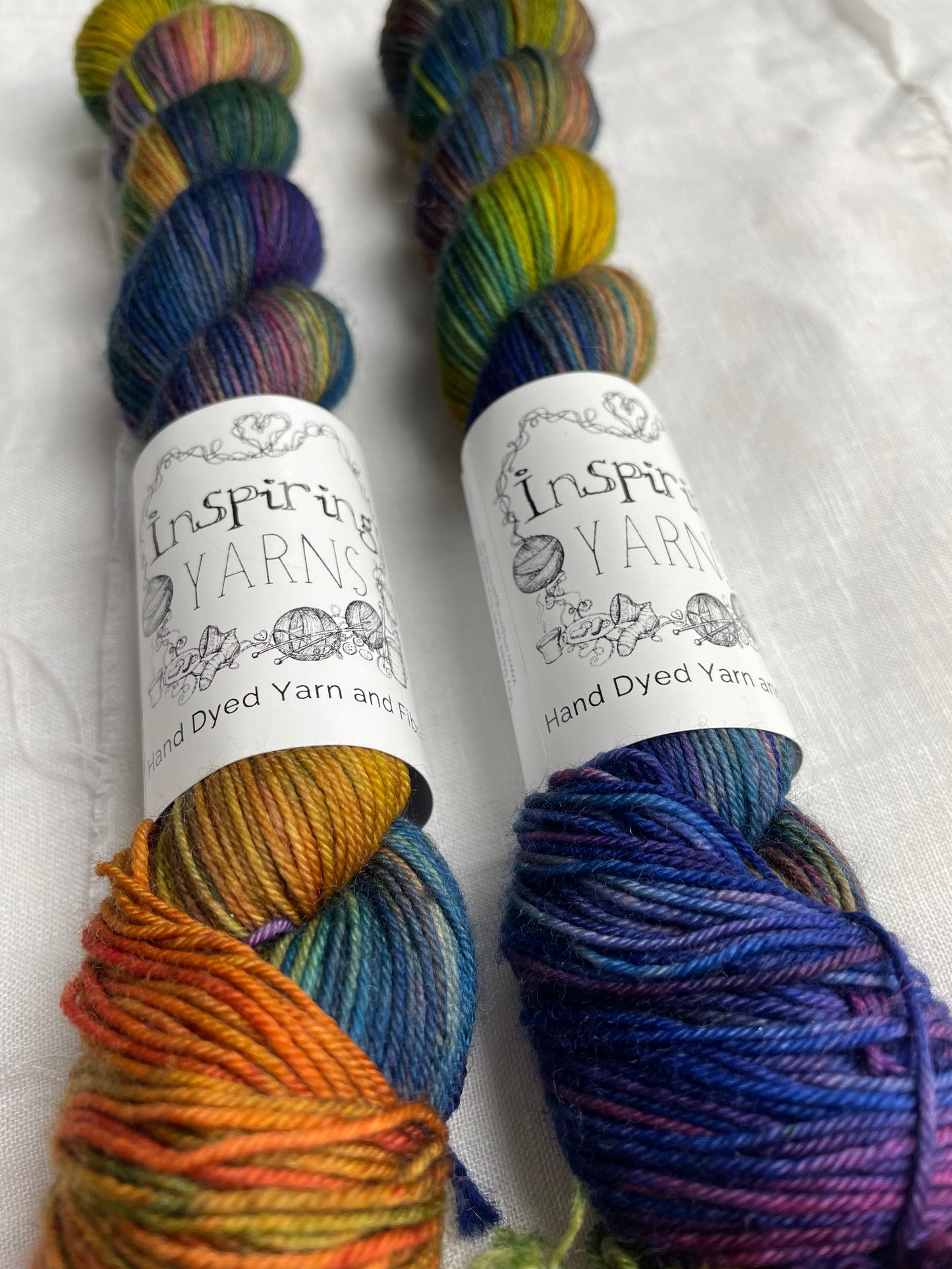Close up of two skeins of multi coloured Merino Smooth Sock yarn labelled Inspiring Yarns on a white linen background.