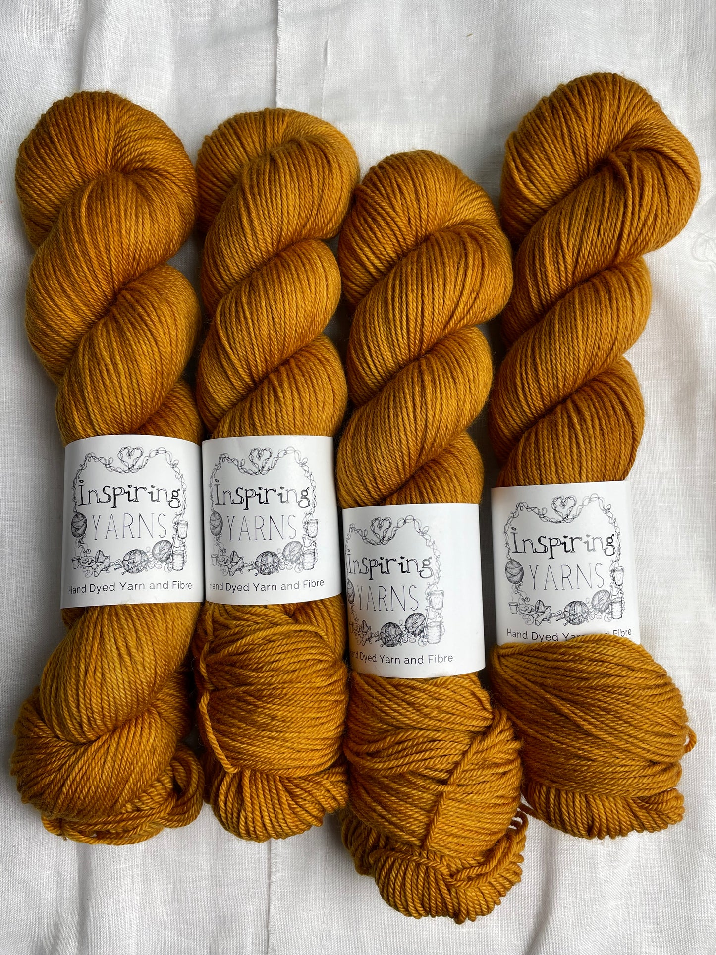 Four skeins of mustard Merino Smooth DK yarn labelled Inspiring Yarns on a white linen background.