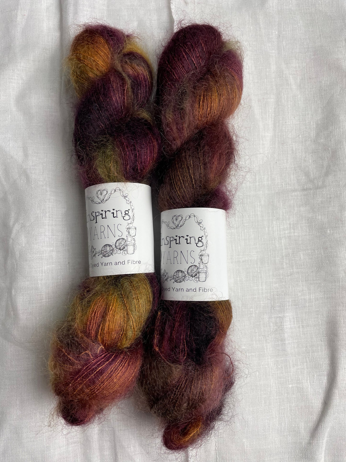 Two skeins of variegated tan burgundy and green mohair silk lace yarn labelled Inspiring Yarns on a white linen background.