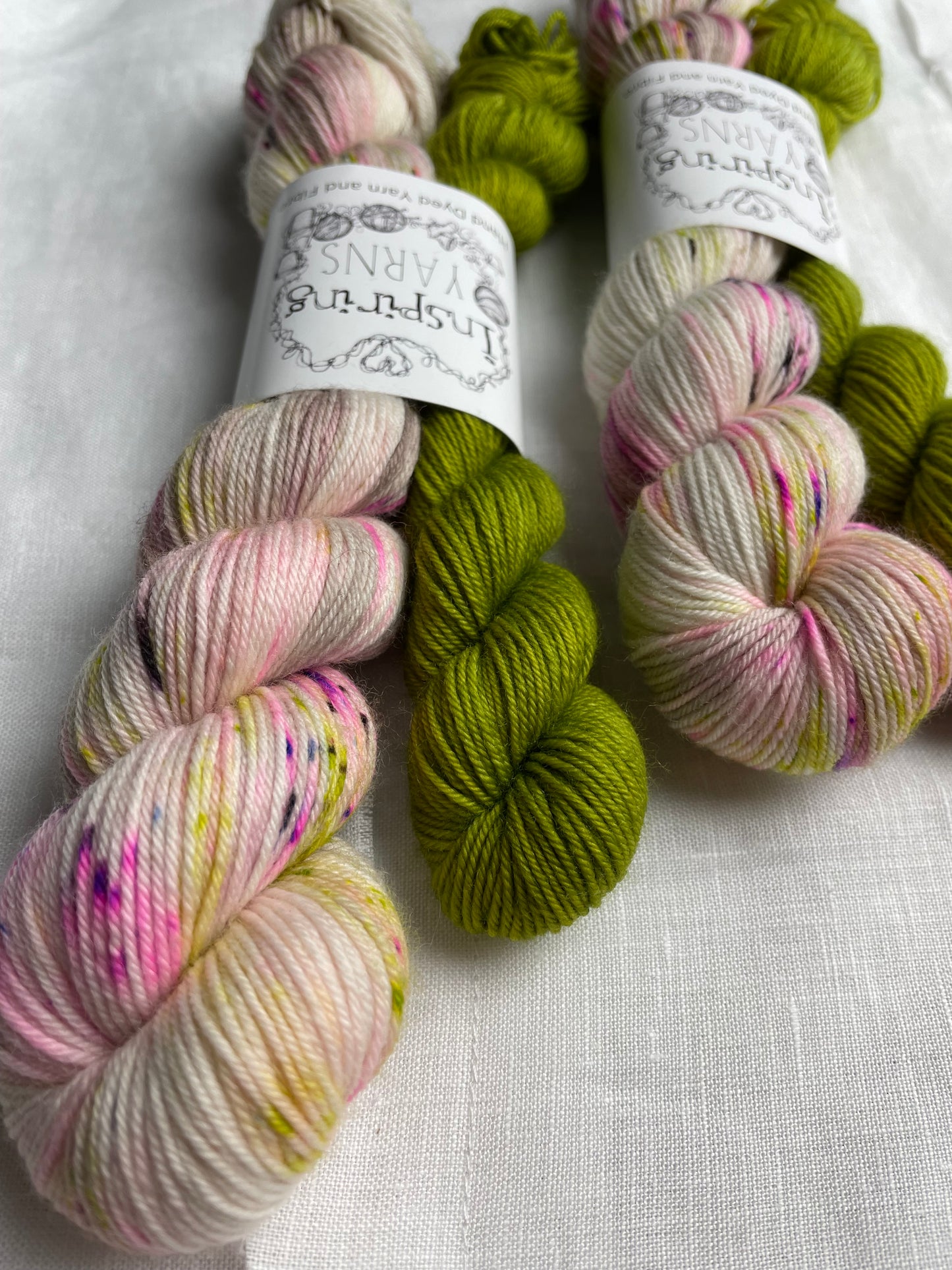 Two sets of hand dyed sock yarn comprising one 50g natural speckled and one 20g bright green on a white linen background. The yarn is branded Inspiring Yarns.