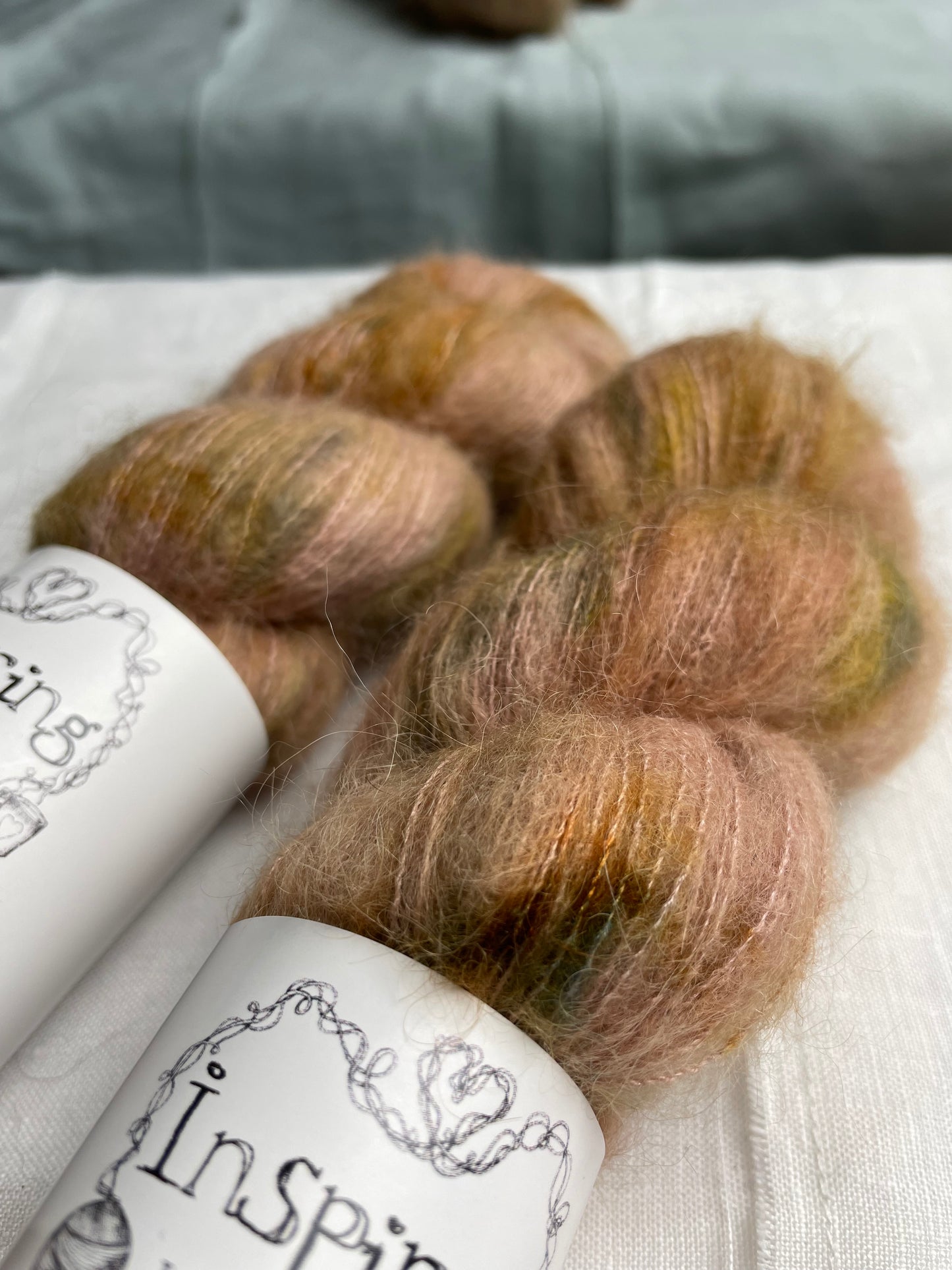 Close up of two skeins of speckled soft terracotta hand dyed fluffy Alpaca Silk Lace yarn labelled Inspiring Yarns on a white linen background.