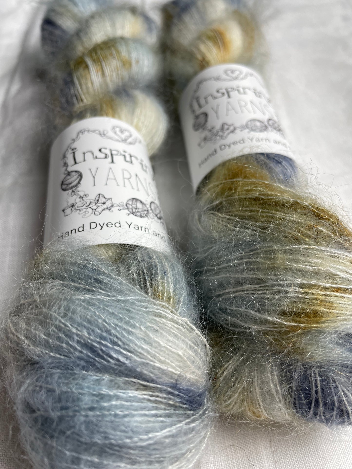 Two skeins of blue and brown speckled mohair silk lace yarn labelled Inspiring Yarns on a white linen background.