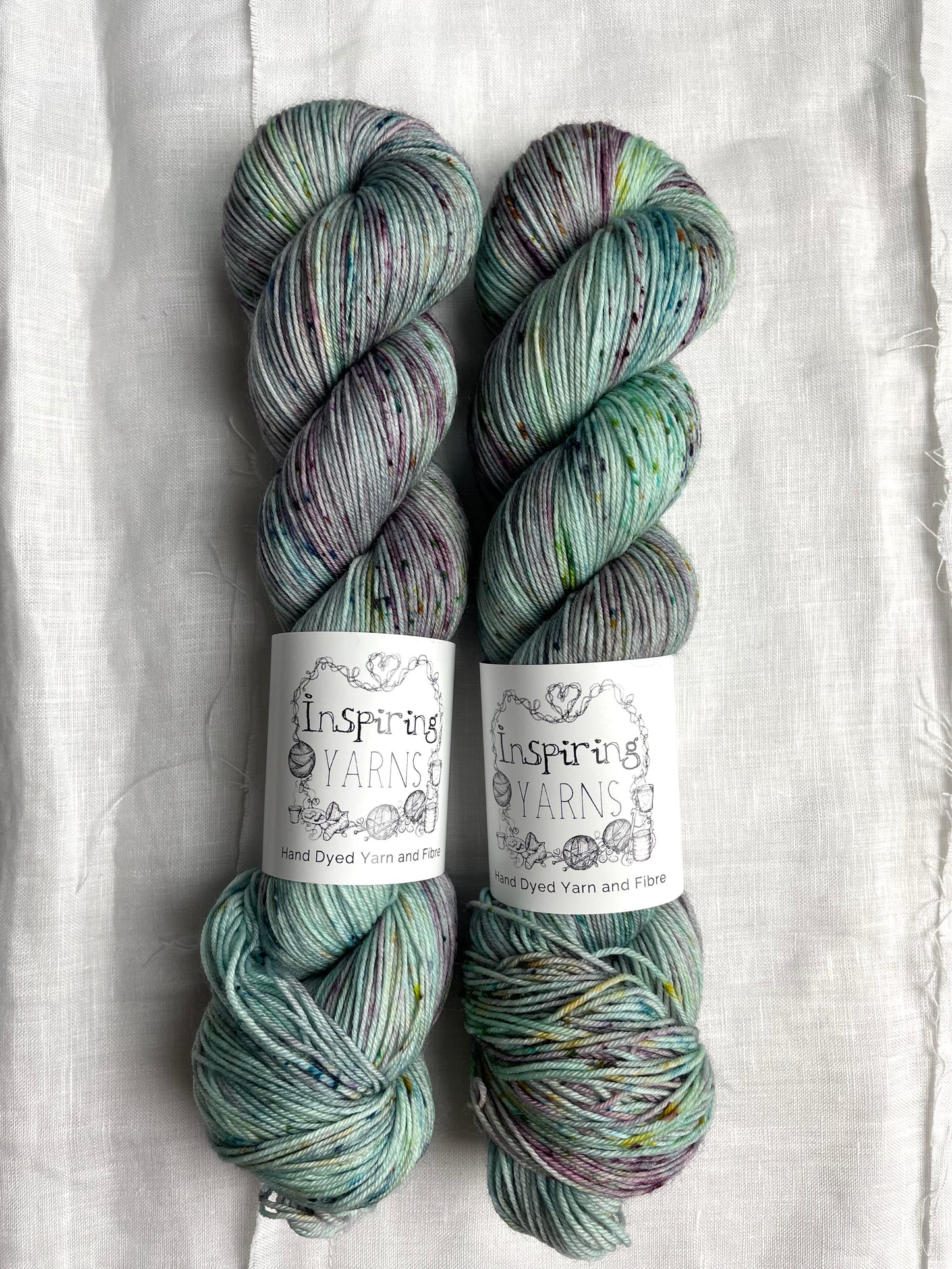 Two skeins of merino smooth sock yarn in a light teal colour with coloured speckles labelled Inspiring Yarns on a white linen background.