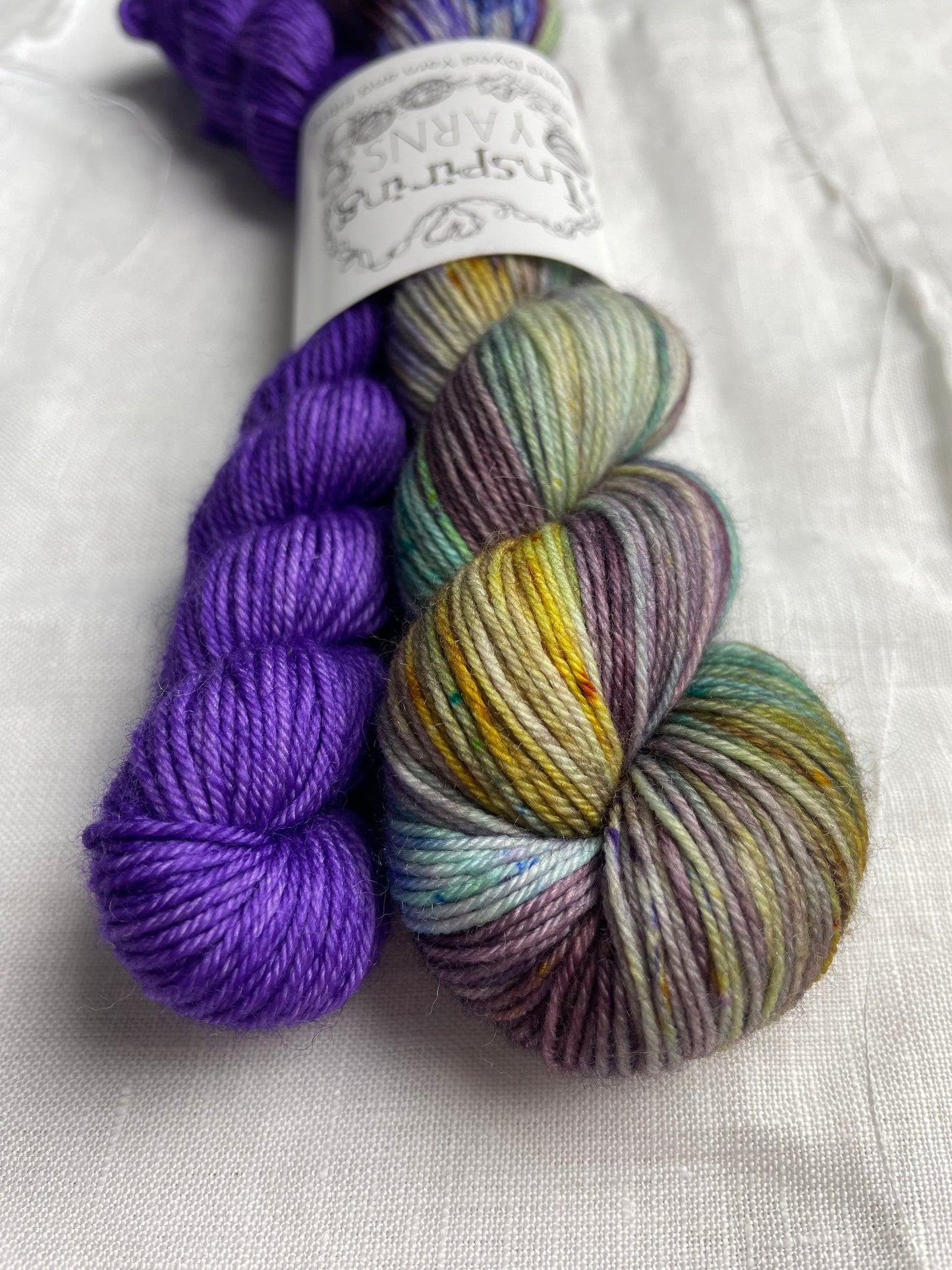 Close up of a set of hand dyed sock yarn comprising one 50g dark plum speckled and one 20g bright purple on a white linen background. The yarn is branded Inspiring Yarns.