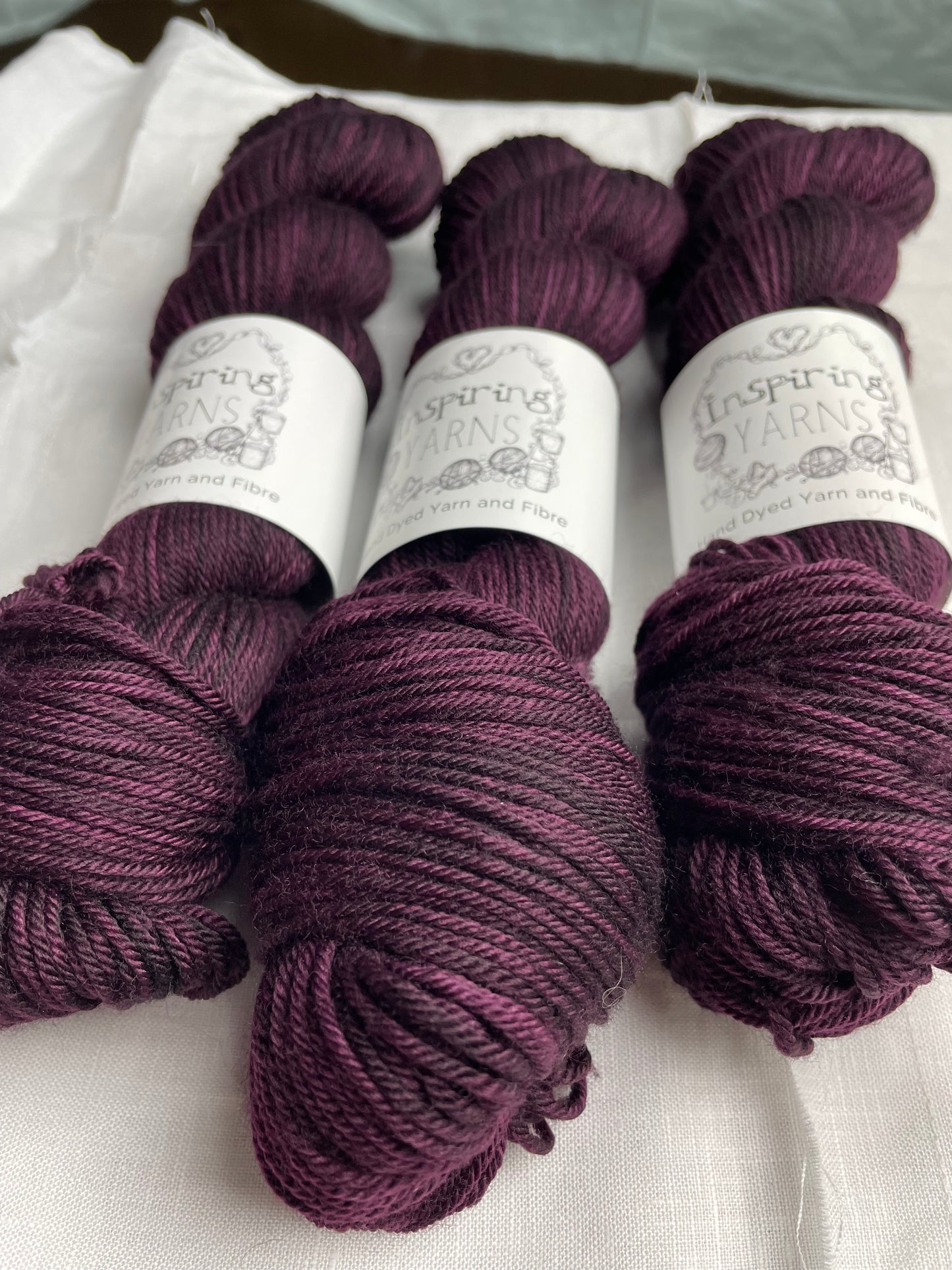 Three skeins of merino smooth DK yarn in a tonal dark burgundy colour labelled Inspiring Yarns on a white linen background.