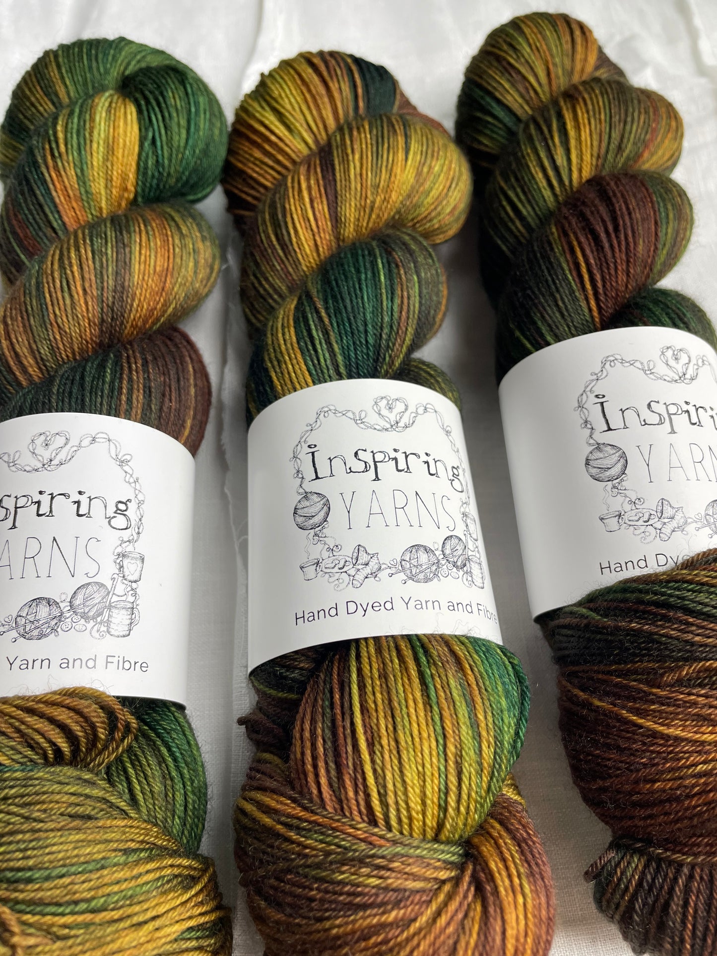 up close of three skeins of yarn with goldens yellows, greens and browns with black and white inspiring yarns labels.