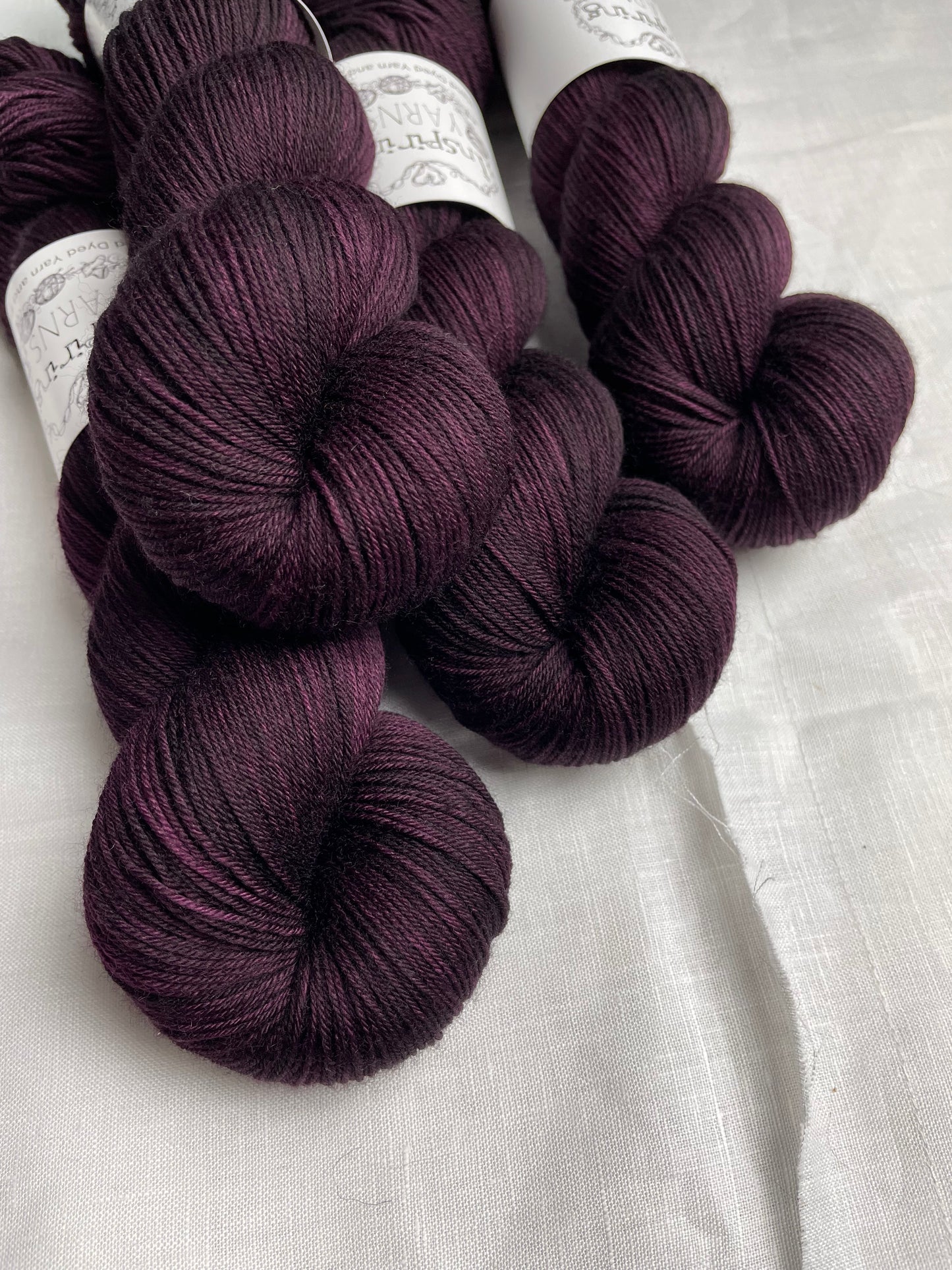 Four skeins of merino smooth sock yarn in a tonal dark burgundy colour labelled Inspiring Yarns on a white linen background.