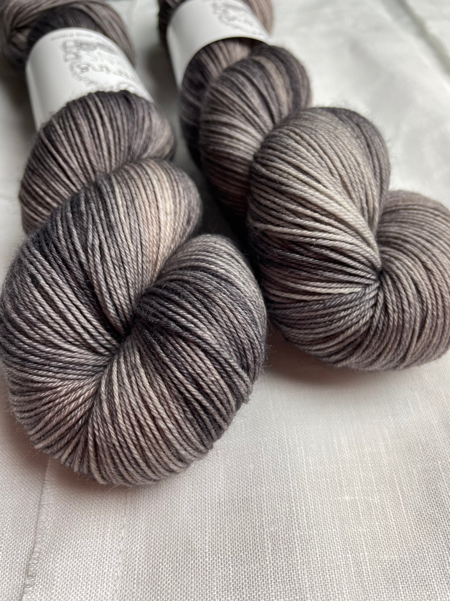 Two skeins of Merino Smooth Sock yarn hand dyed in a tonal grey colourway and labelled Inspiring Yarns on a white linen background.