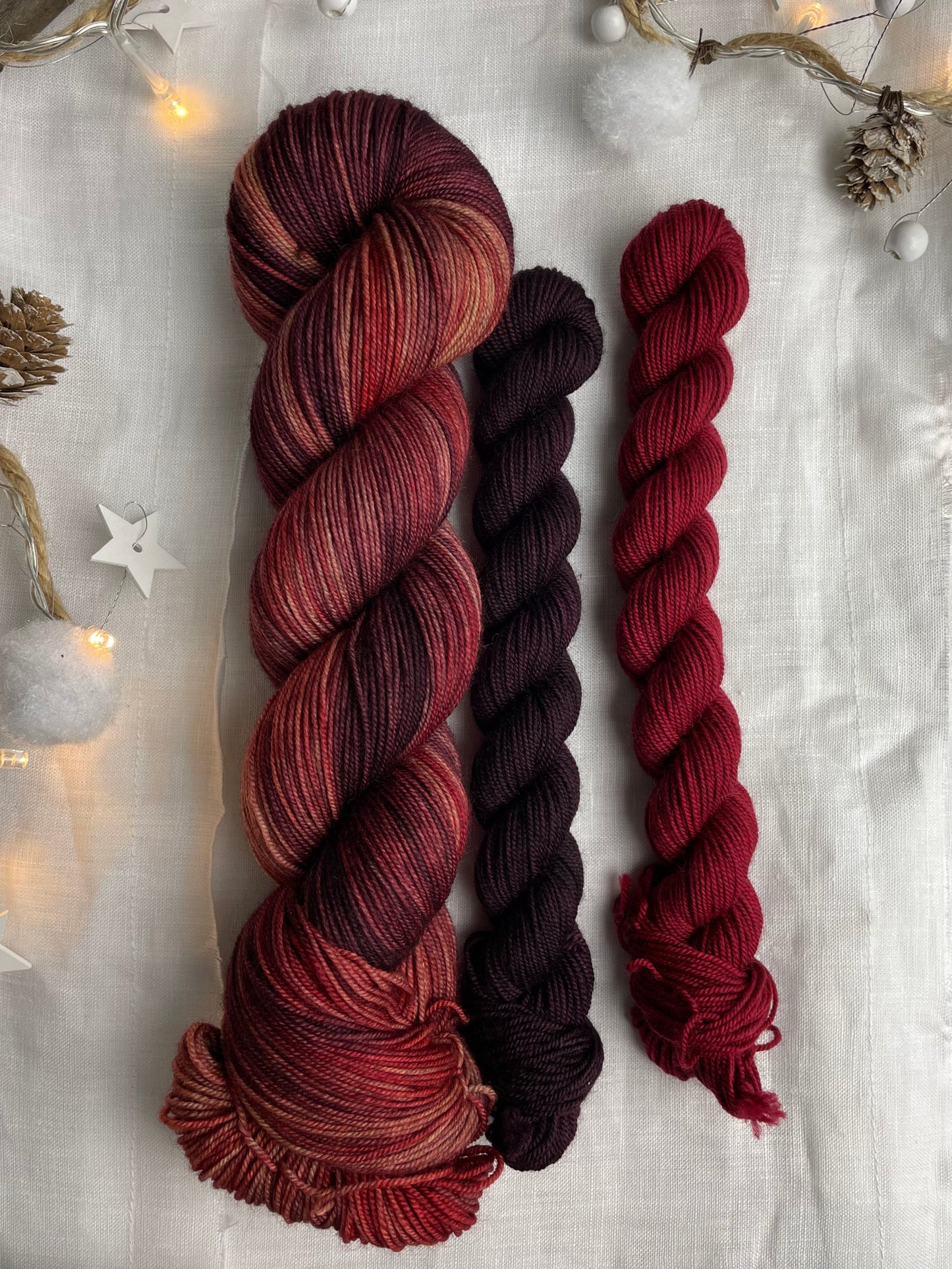 A hand dyed yarn sock set featuring large skein in burgundy, red and orange tones with a dark red and a burgundy mini skein labelled Inspiring Yarns. The background is white linen and some Christmas lights with pinecones, twigs and stars.
