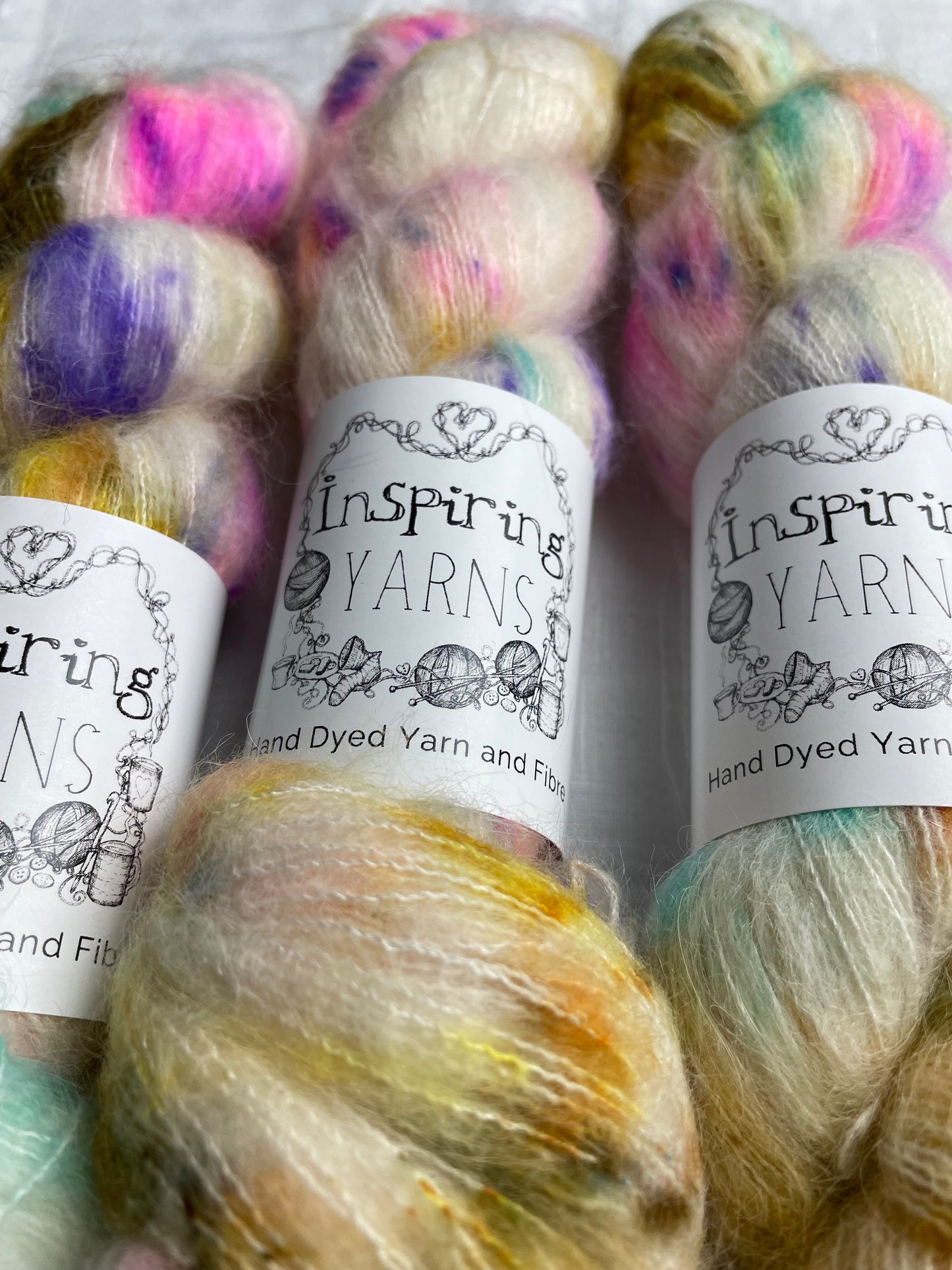 Close up of three skeins of brightly speckled natural coloured hand dyed fluffy Alpaca Silk Lace yarn labelled Inspiring Yarns on a white linen background.