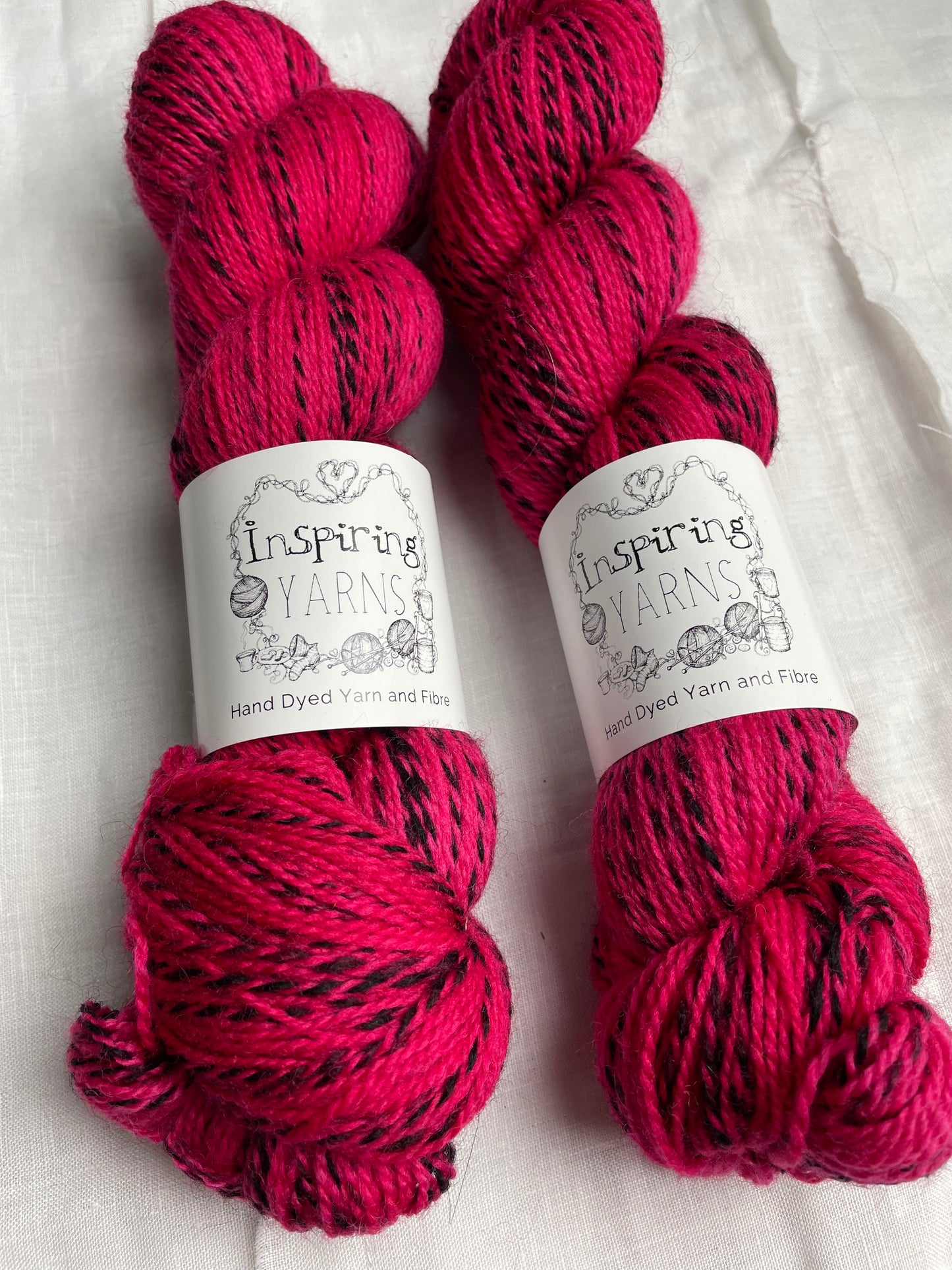 Close up of two skeins of handyed Highland Zebra yarn in a bright cerise pink colourway. The yarn has black strands spun in to it. The skeins are labelled Inspiring Yarns.