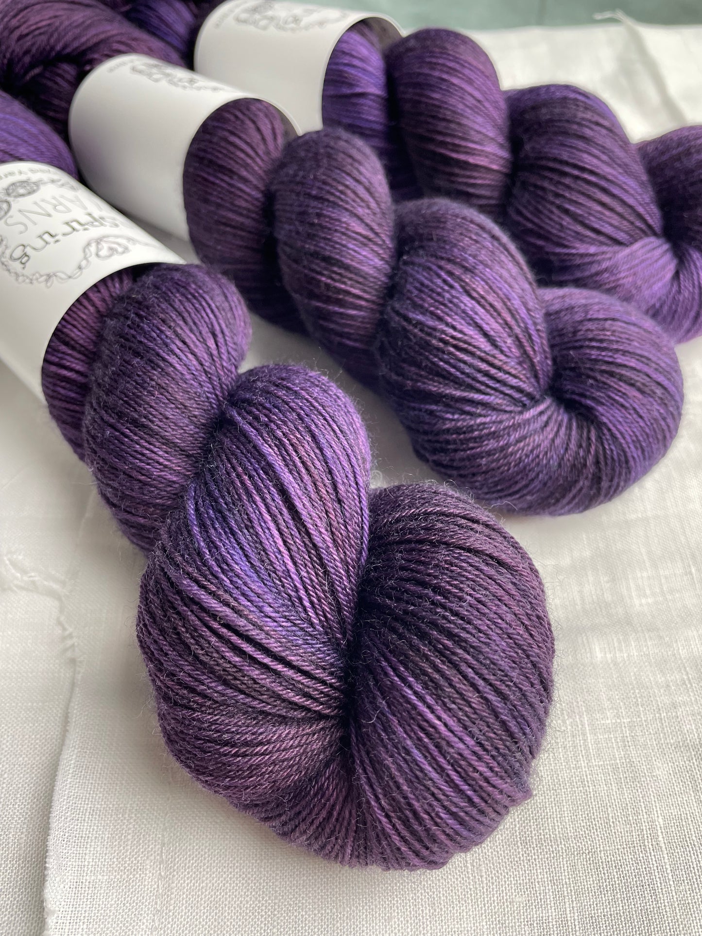 Three skeins of merino smooth sock yarn in a tonal dark purple colour labelled Inspiring Yarns on a white linen background.