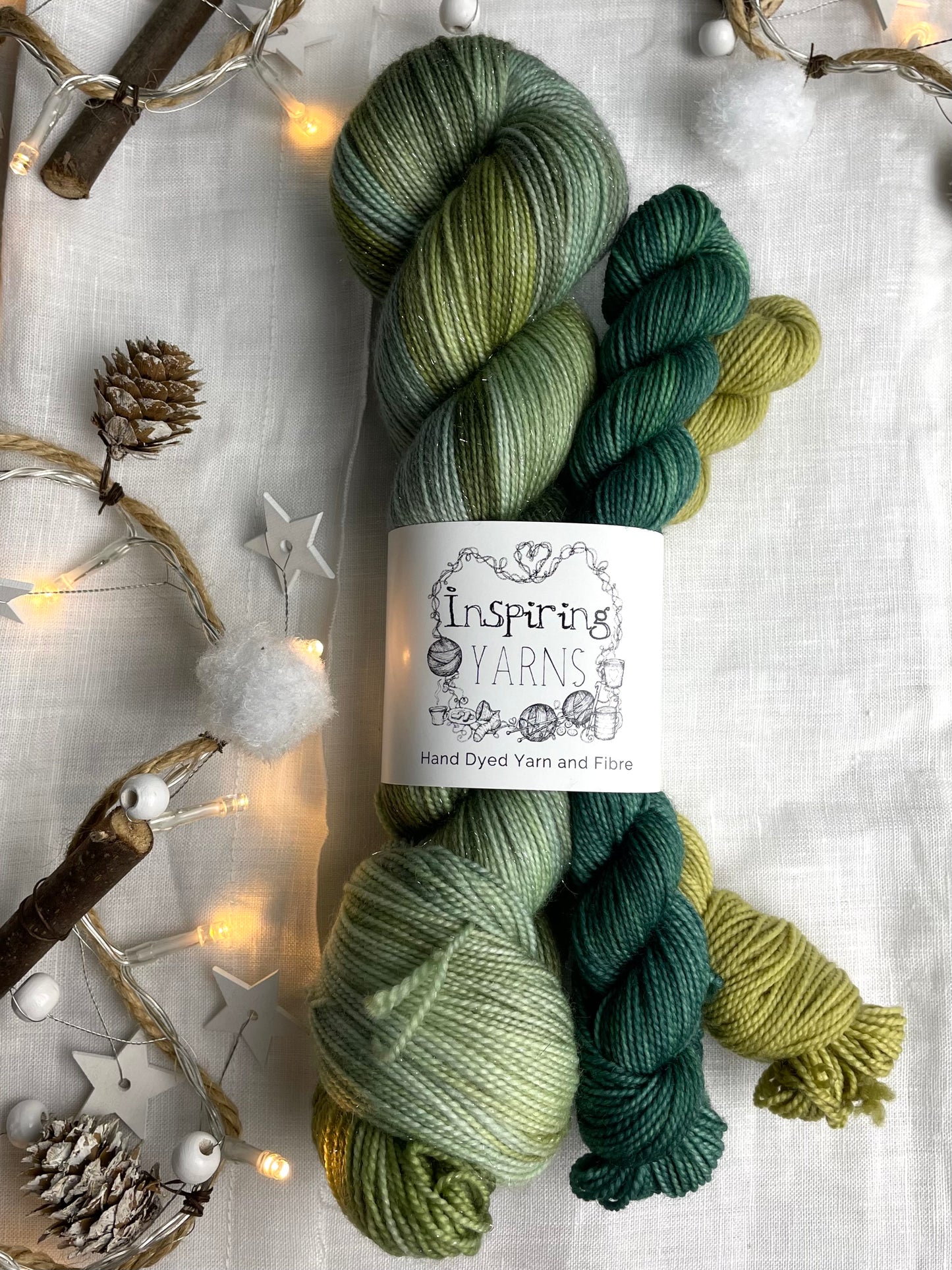 A hand dyed yarn sock set featuring large skein of Sparkle Sock Yarn in green tones with a teal and a lime green mini skein labelled Inspiring Yarns. The background is white linen and some Christmas lights with pinecones, twigs and stars.