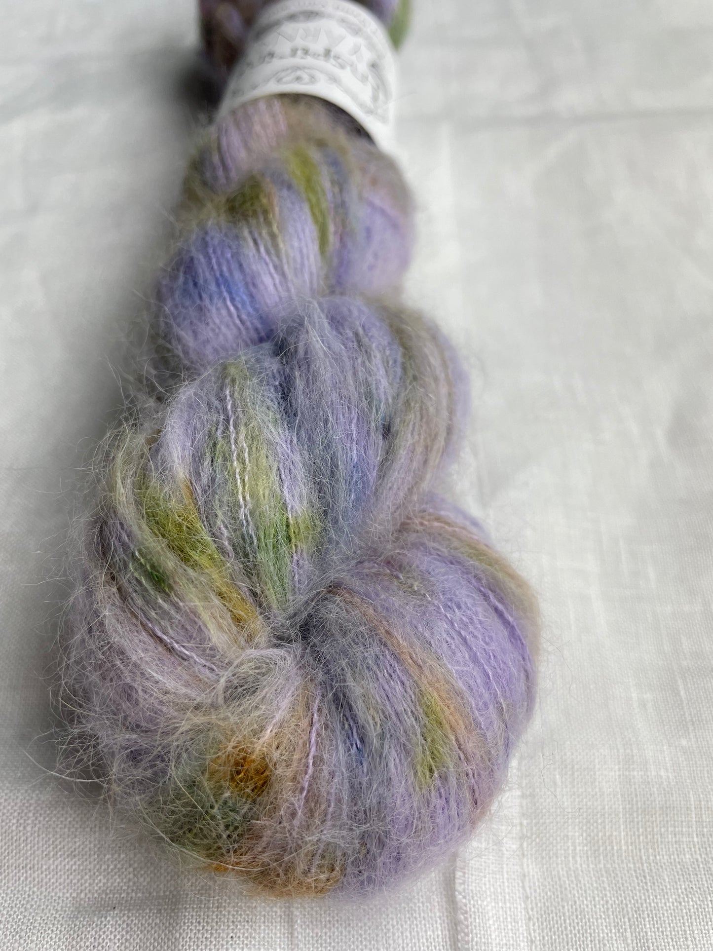 Close up of a skein of speckled lilac coloured hand dyed fluffy Alpaca Silk Lace yarn labelled Inspiring Yarns on a white linen background.