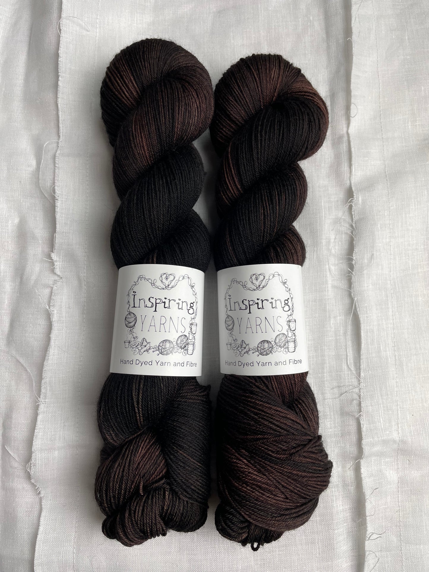 Two skeins of variegated dark brown black Merino Smooth Sock yarn labelled Inspiring Yarns on a white linen background